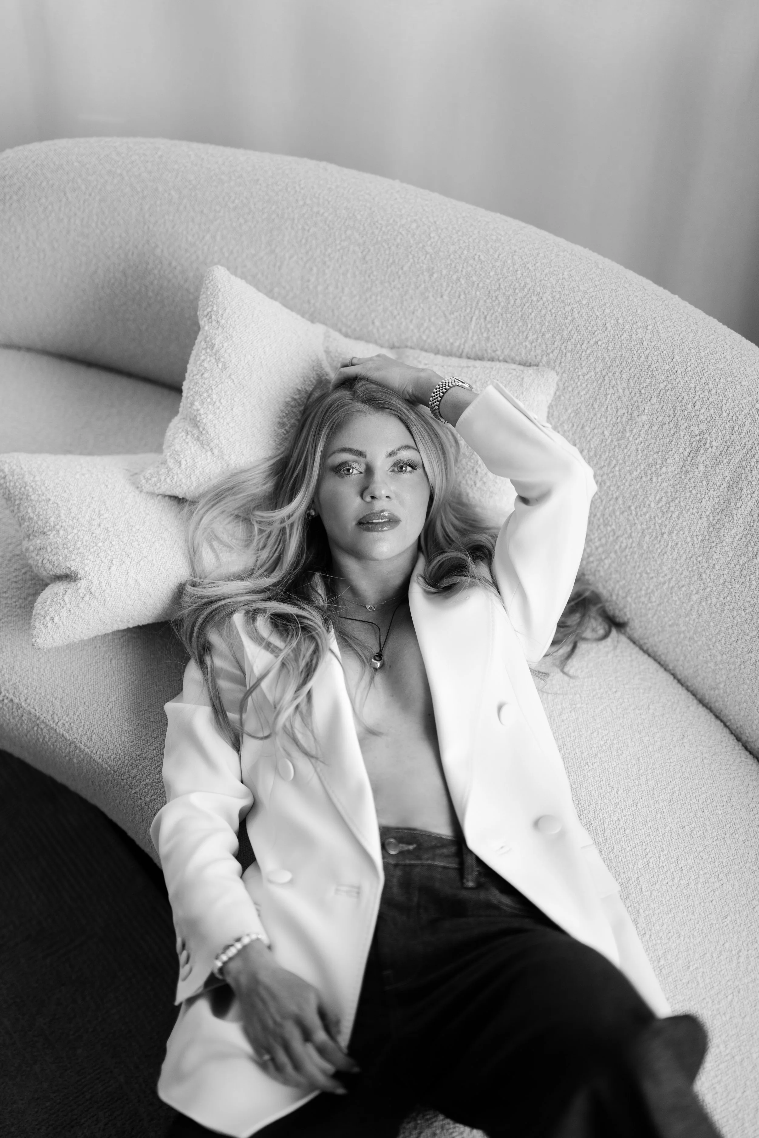 Black and white photo of a woman with long wavy hair lying on a textured sofa, gazing at the camera with her hand resting on her head, wearing an open white blazer and dark pants.