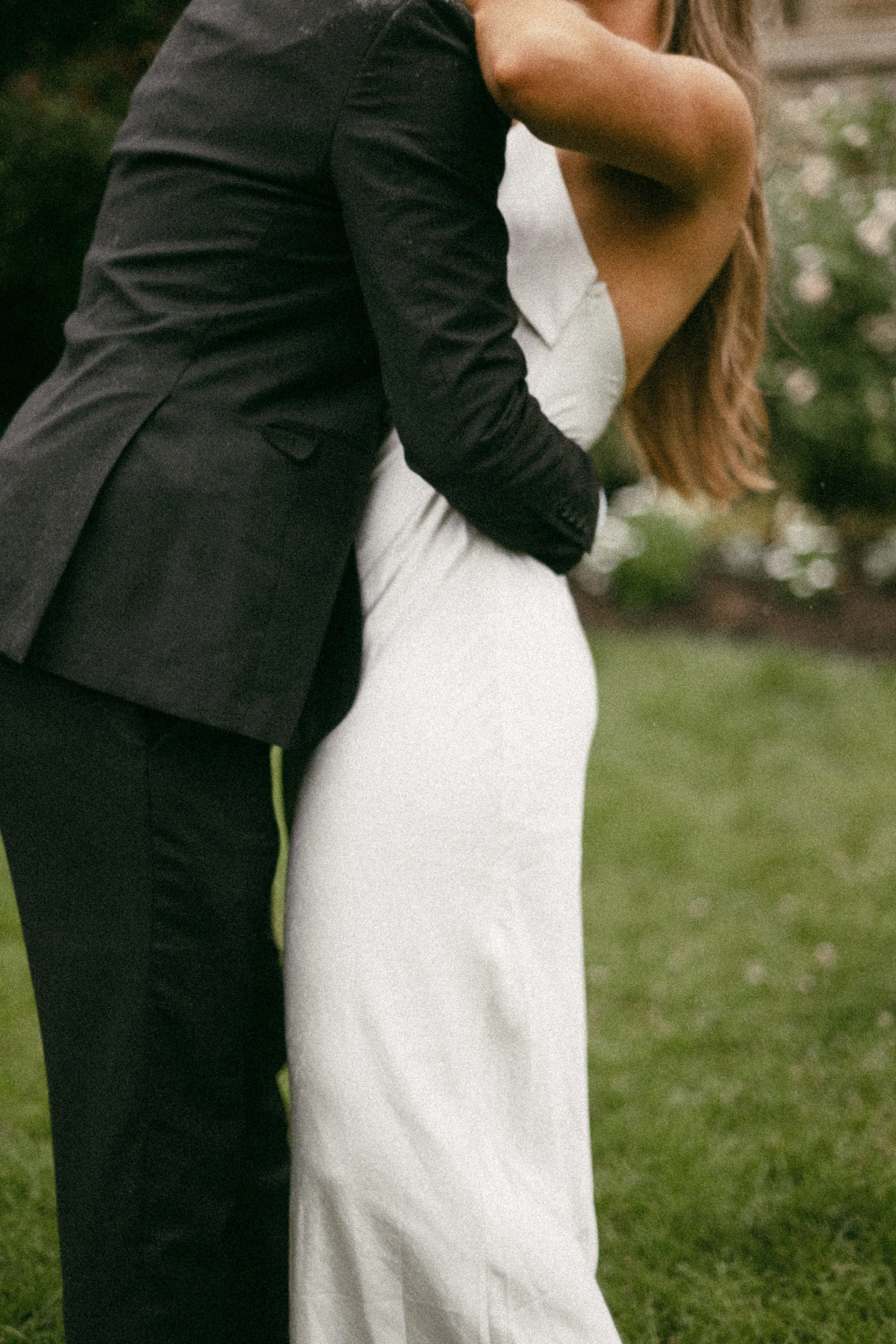 A couple embraces outdoors, with the man wearing a black suit and the woman in a white dress.