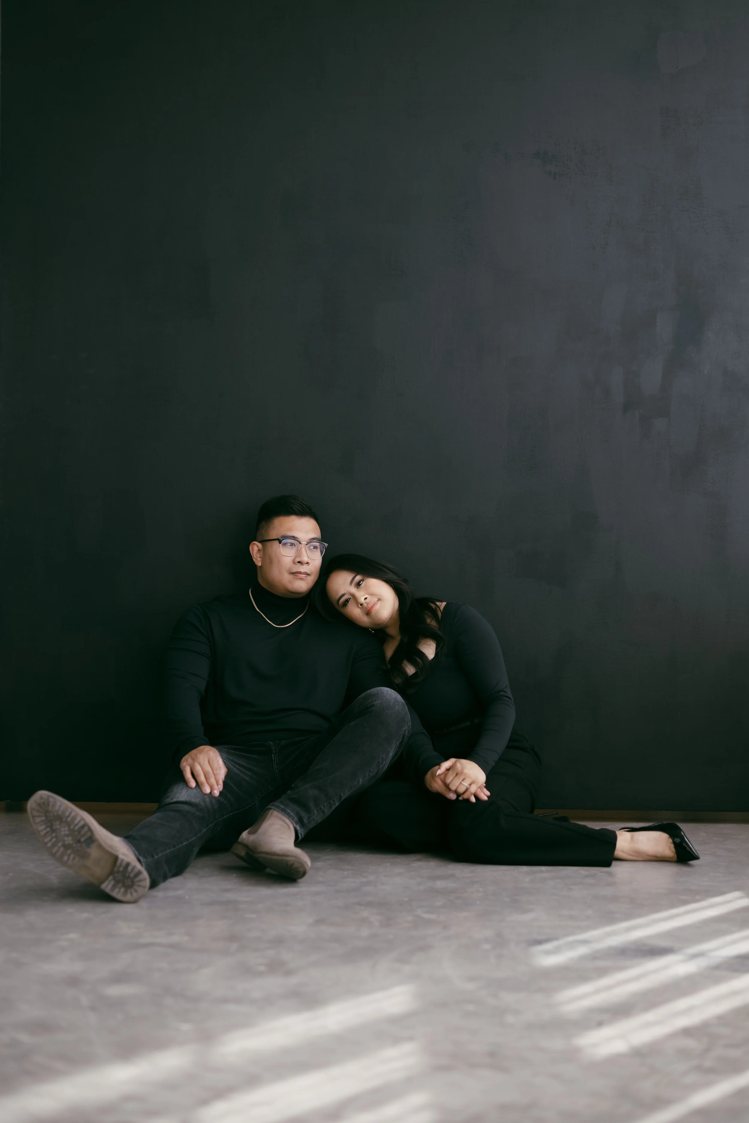 A man and woman sitting on the floor against a dark wall, with the woman resting her head on the man's shoulder, both dressed in black.