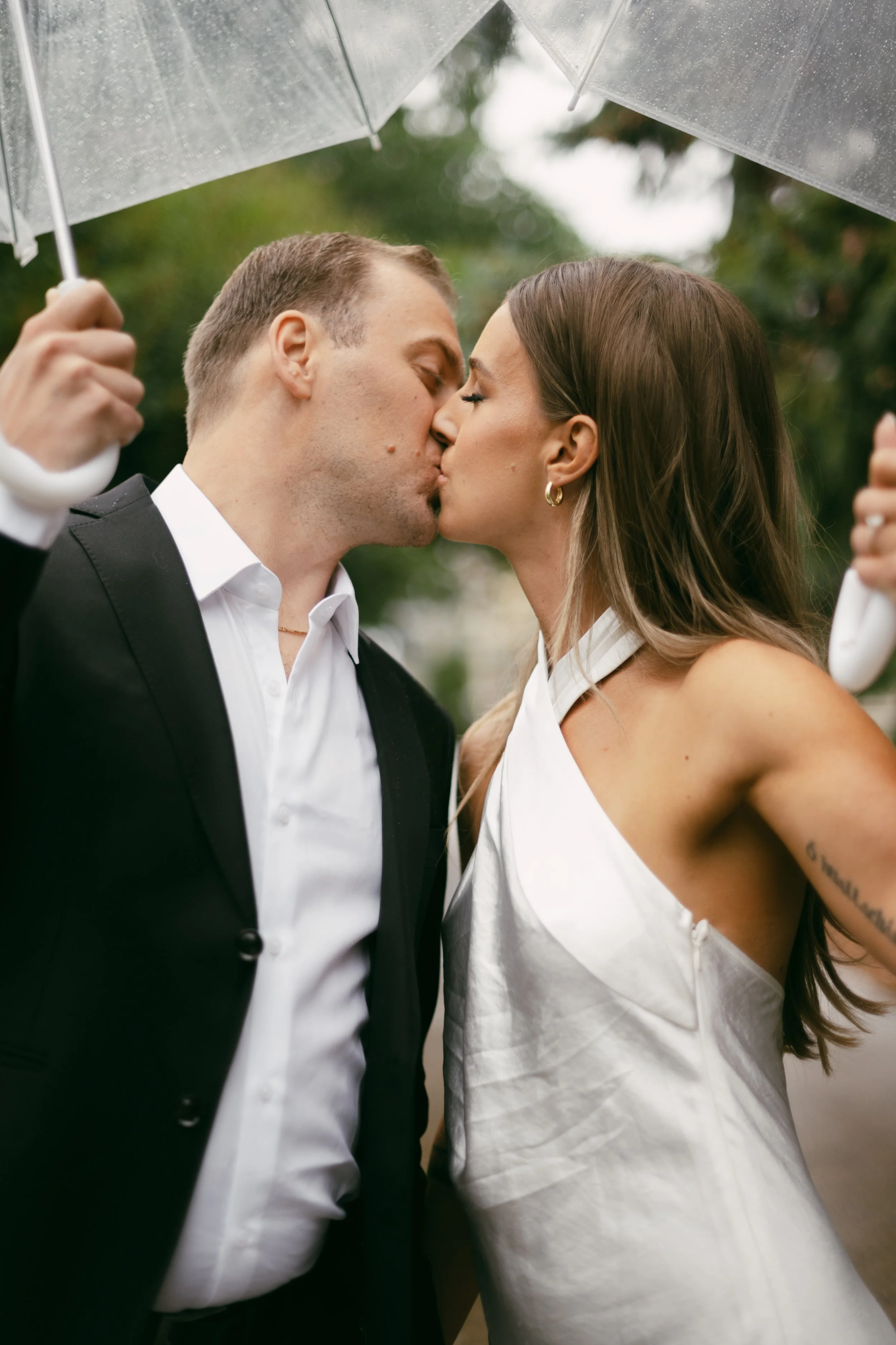 A couple dressed in formal attire sharing a kiss under a transparent umbrella outdoors.