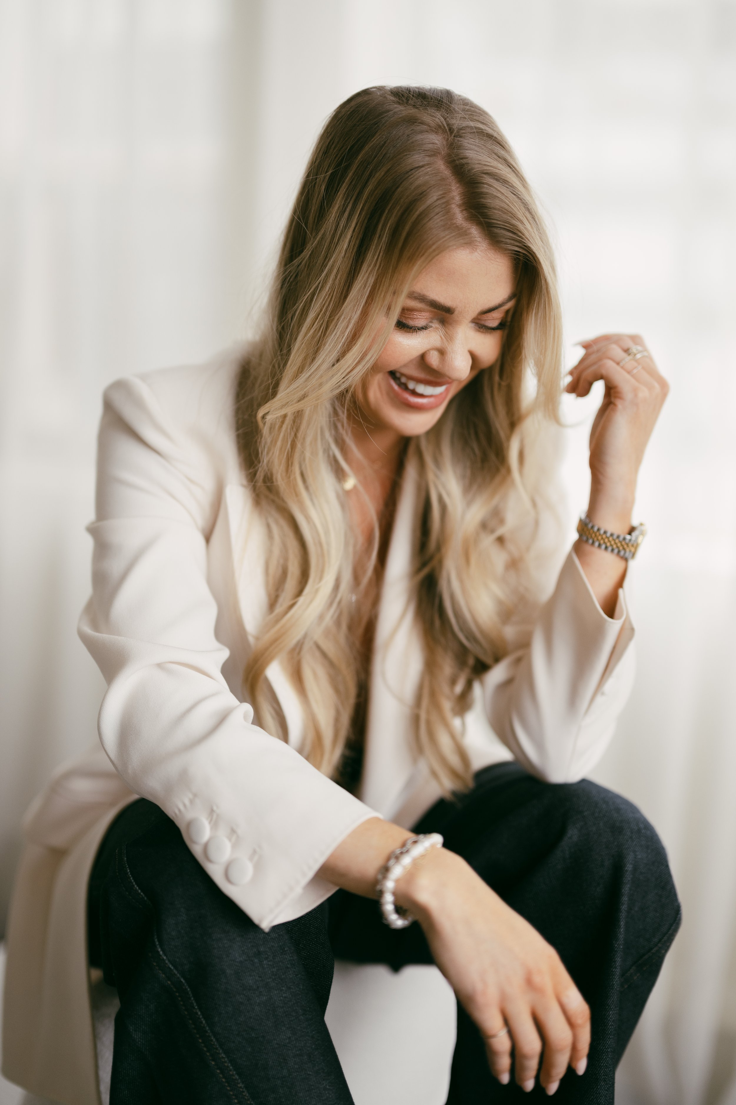 A woman with long blonde hair wearing a white blazer and black pants, sitting and smiling with her eyes closed.