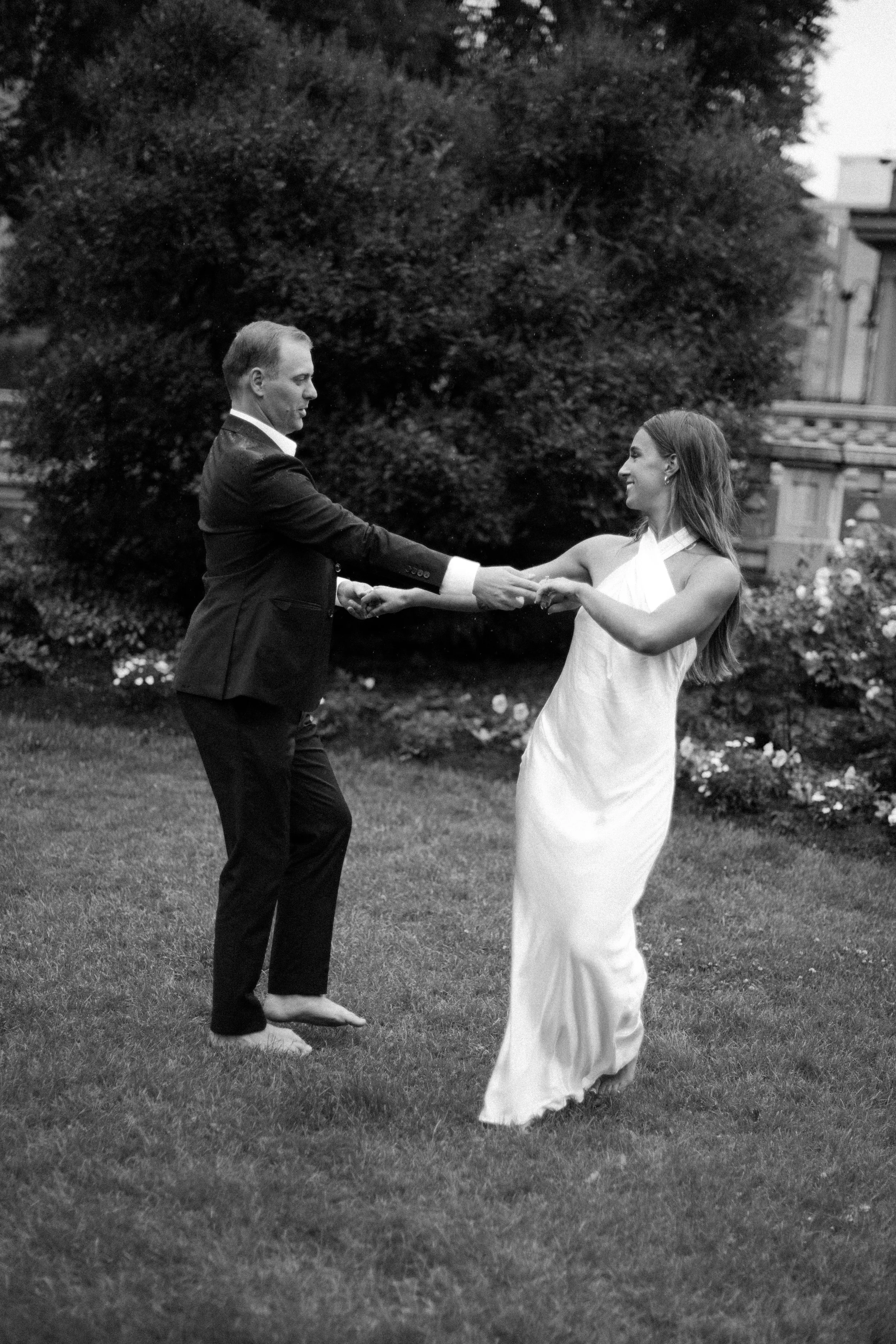 A black and white photo of a couple dancing outdoors. The man is in a suit and the woman in a long dress, holding hands and smiling.