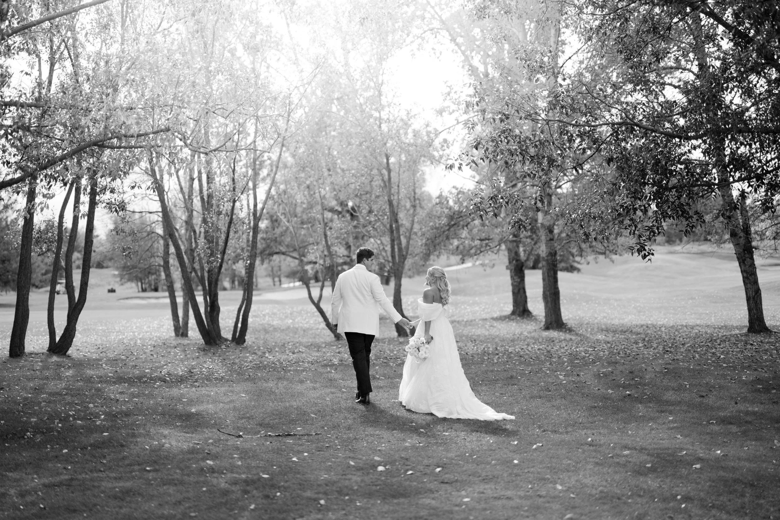 A black and white photo of a bride and groom walking hand in hand through a park with trees, sunlight shining down, and fallen leaves on the ground.