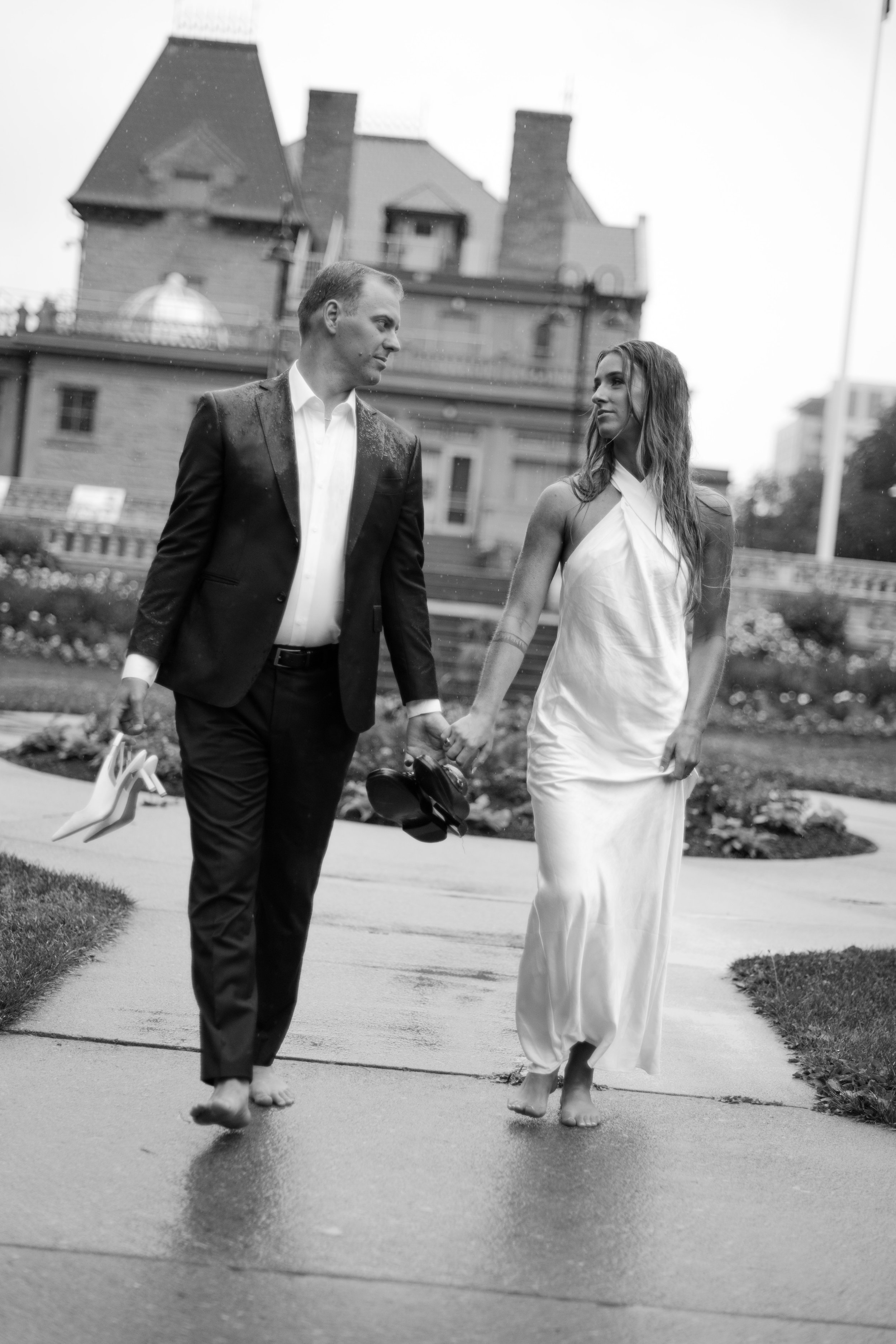 A black and white photo of a man and woman walking barefoot on a sidewalk, holding hands. The man is dressed in a suit and is carrying high-heeled shoes, while the woman is wearing a long white dress and holding her shoes. They are outdoors with a la
