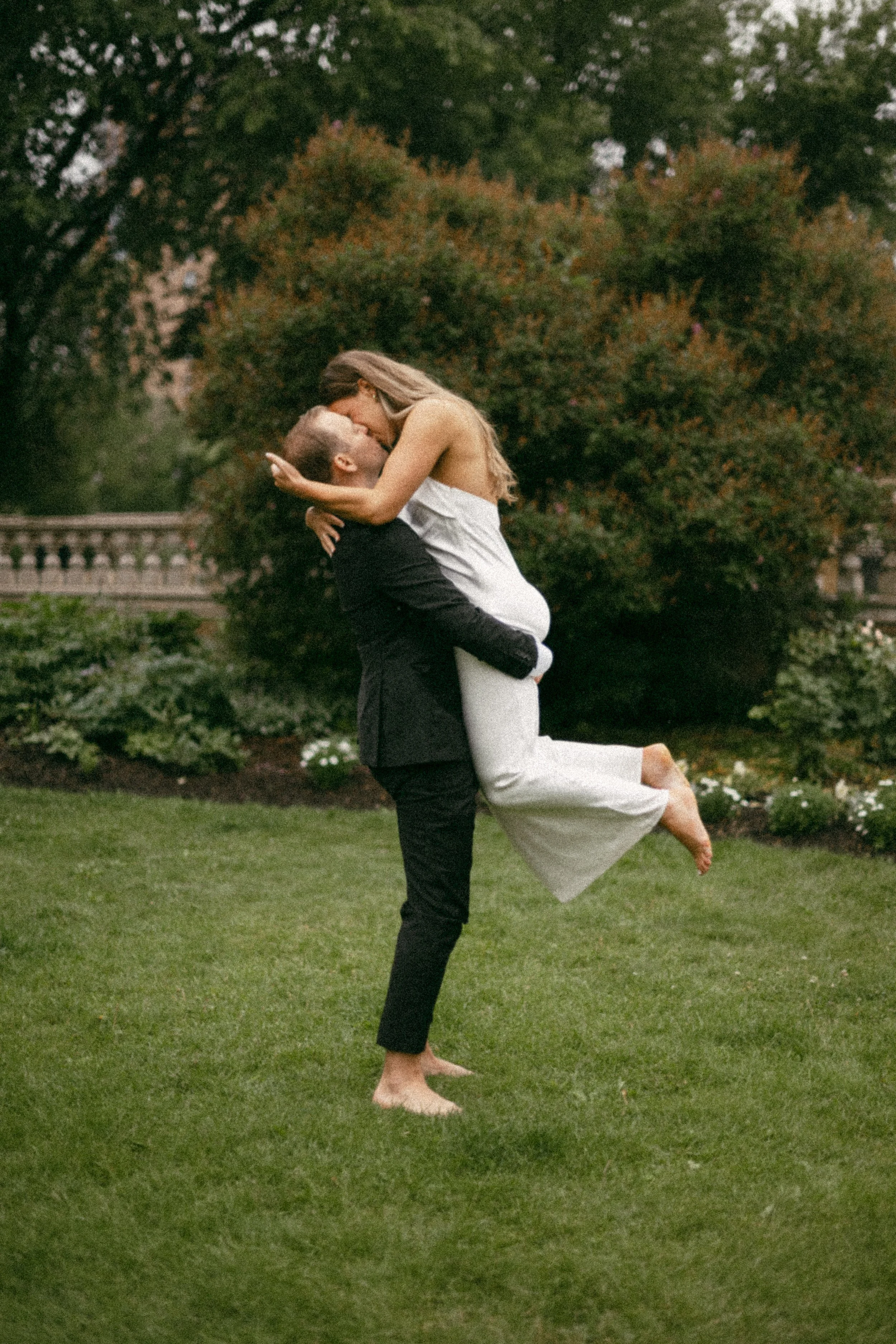 A man in a black suit lifting a woman in a white dress in a garden with trees and bushes, sharing a kiss.