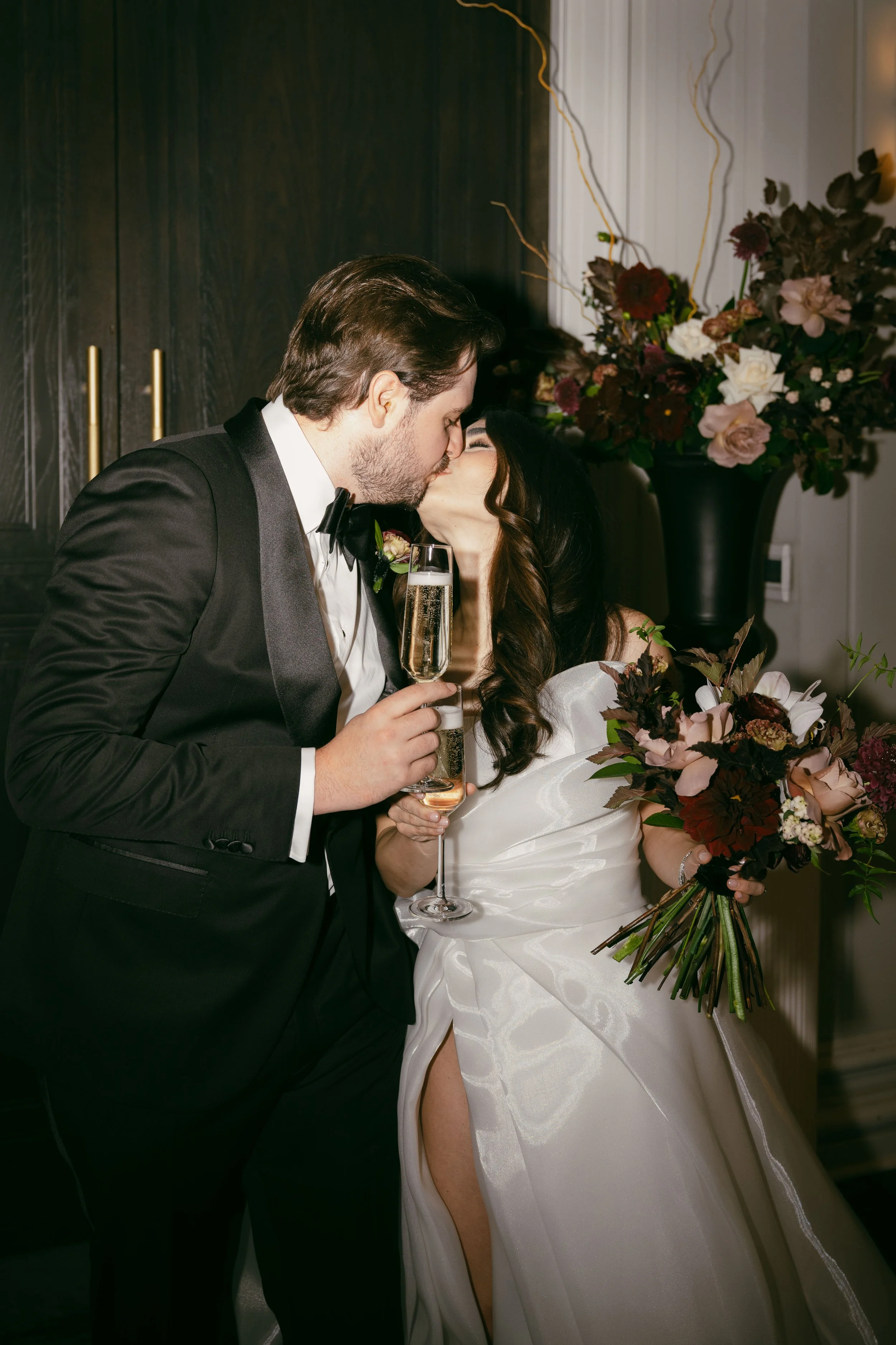 A newlywed couple sharing a kiss on their wedding day, holding champagne glasses. The groom is in a tuxedo, and the bride is in a white gown holding a bouquet of flowers, with a large floral arrangement in the background.