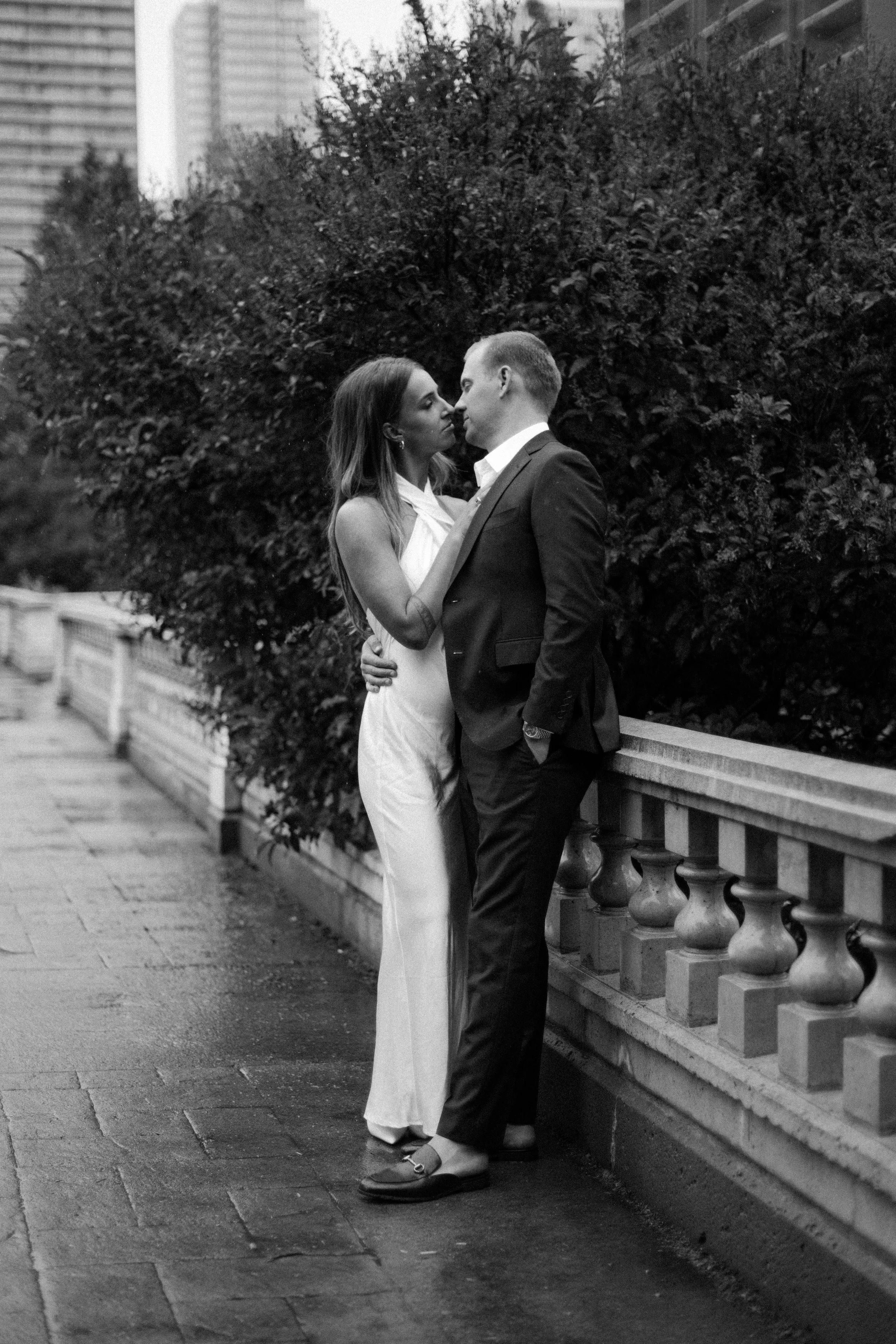 A black and white photo of a man and woman standing close together on a city sidewalk, gazing at each other affectionately. The woman is wearing a sleeveless dress, and the man is dressed in a suit. They are beside a railing with large bushes behind 