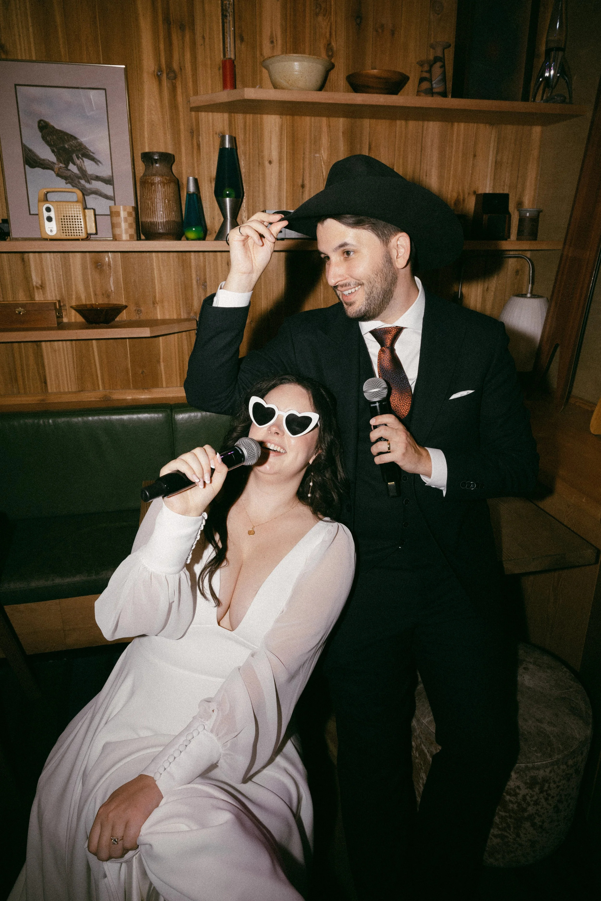 A woman in a white dress and heart-shaped sunglasses sitting on a chair, holding a microphone, with a man in a dark suit and cowboy hat standing next to her, holding a microphone and smiling indoors.