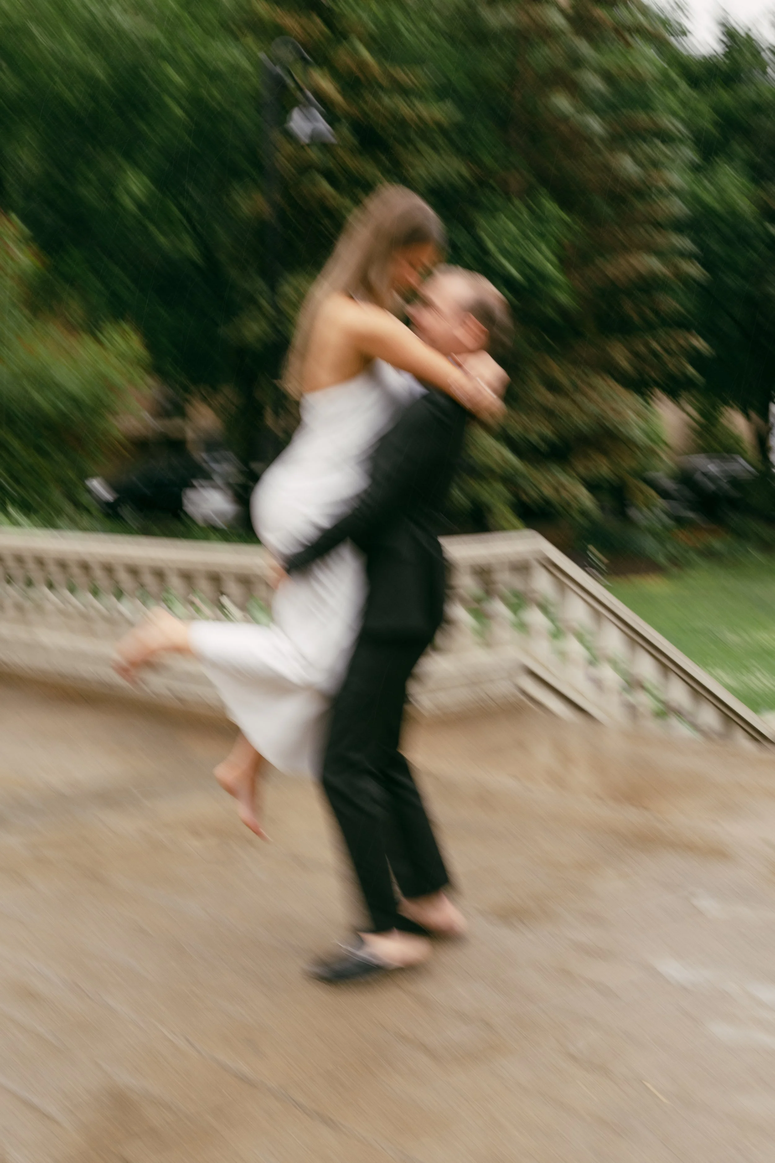 A couple dressed in formal attire, the woman in a white dress and the man in a black suit, is outdoors on a staircase, with the woman riding on the man's shoulders during daytime. The movement creates a blurry effect.