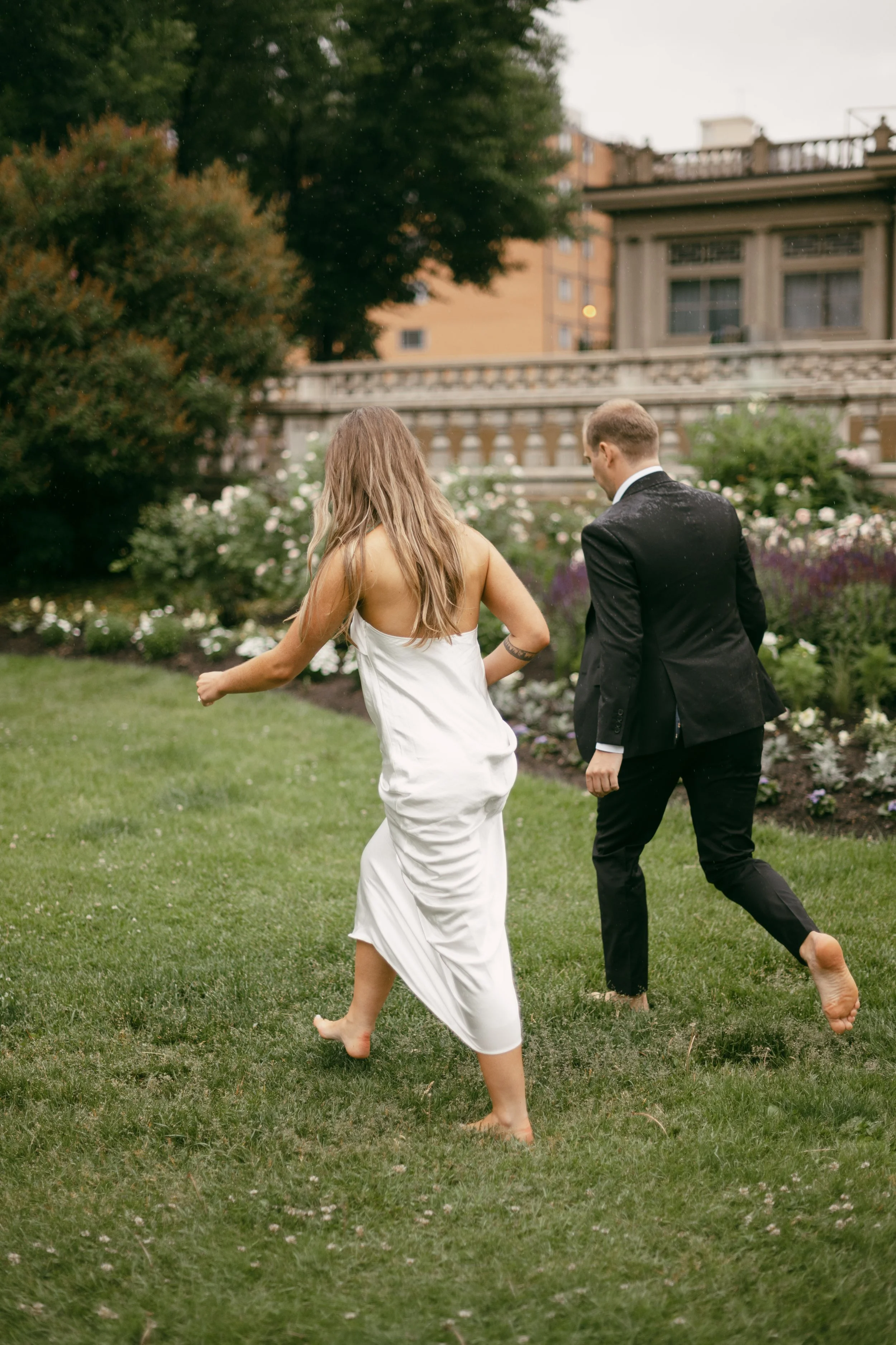 A woman in a white dress and a man in a black tuxedo are running barefoot in a garden with flower beds and greenery.