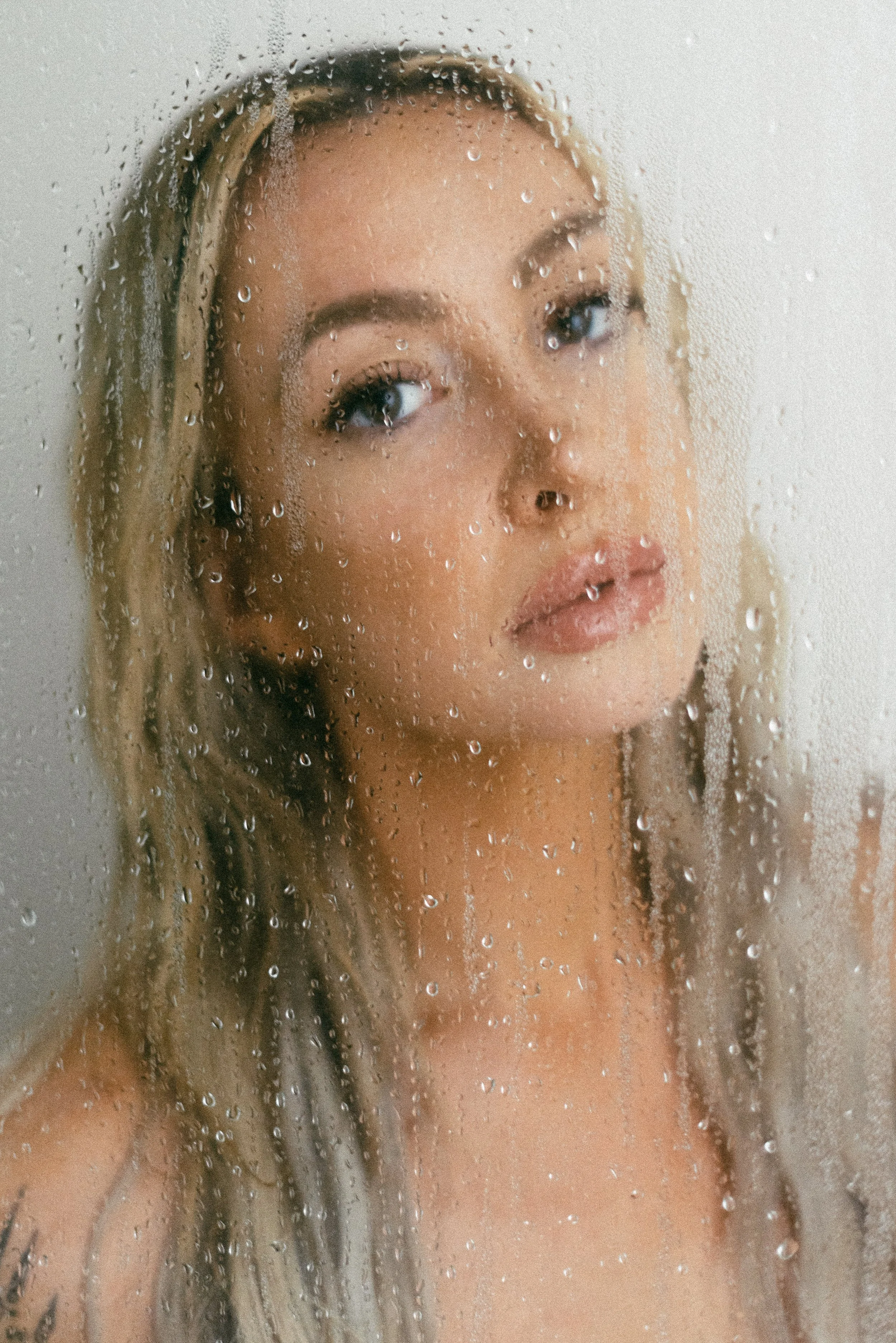 A woman with blonde hair looking through a foggy or rain-covered glass surface.