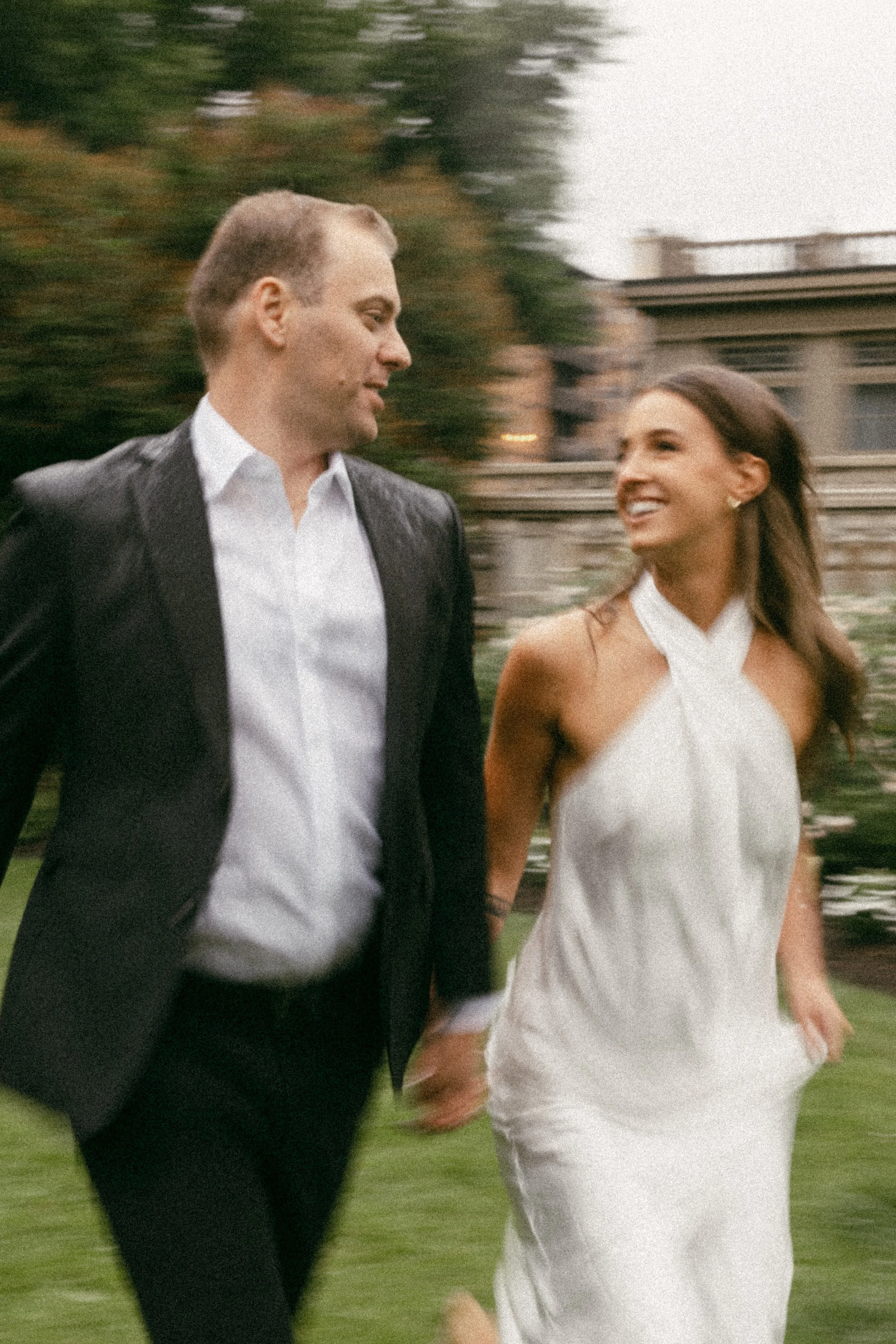 A man and woman holding hands and walking outdoors, smiling at each other, with a blurred background of a house and trees.