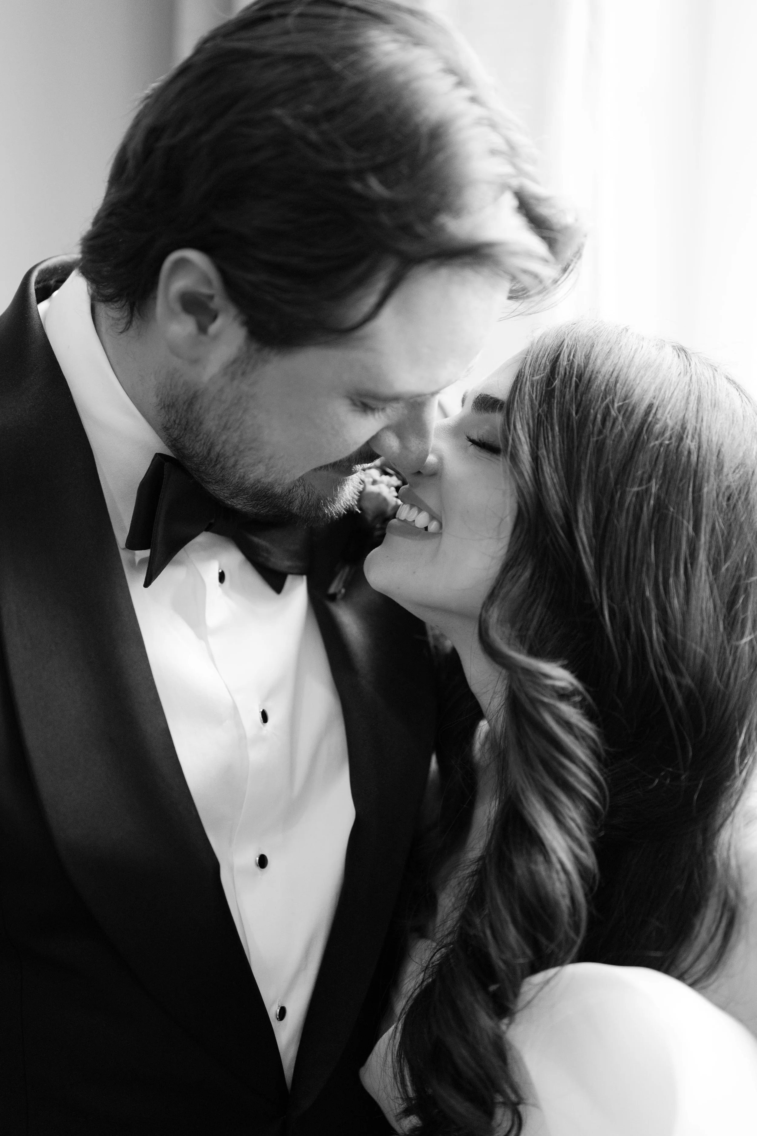 Black and white photo of a couple smiling with their noses touching, dressed in formal attire, close-up shot.