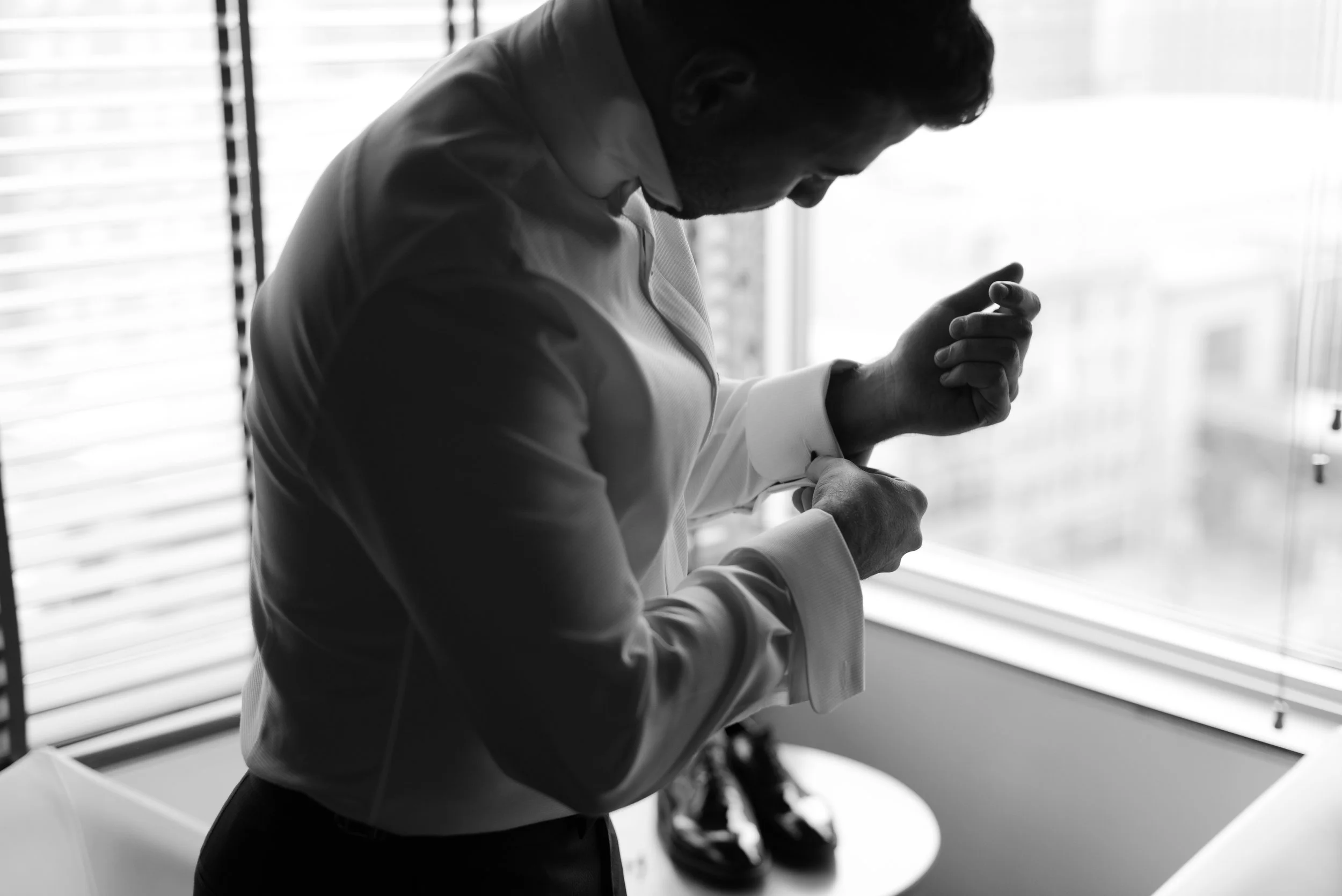Man adjusting cufflinks on his shirt in front of a window.