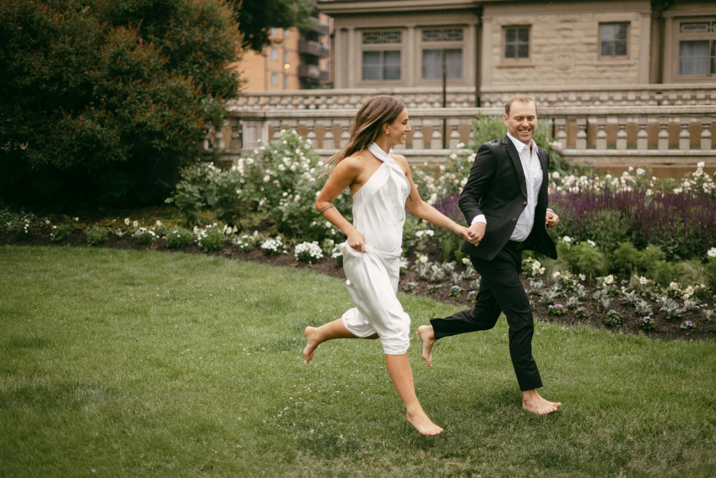 A couple running barefoot on a lawn, holding hands in a garden with flowers and trees, with a historic building in the background.