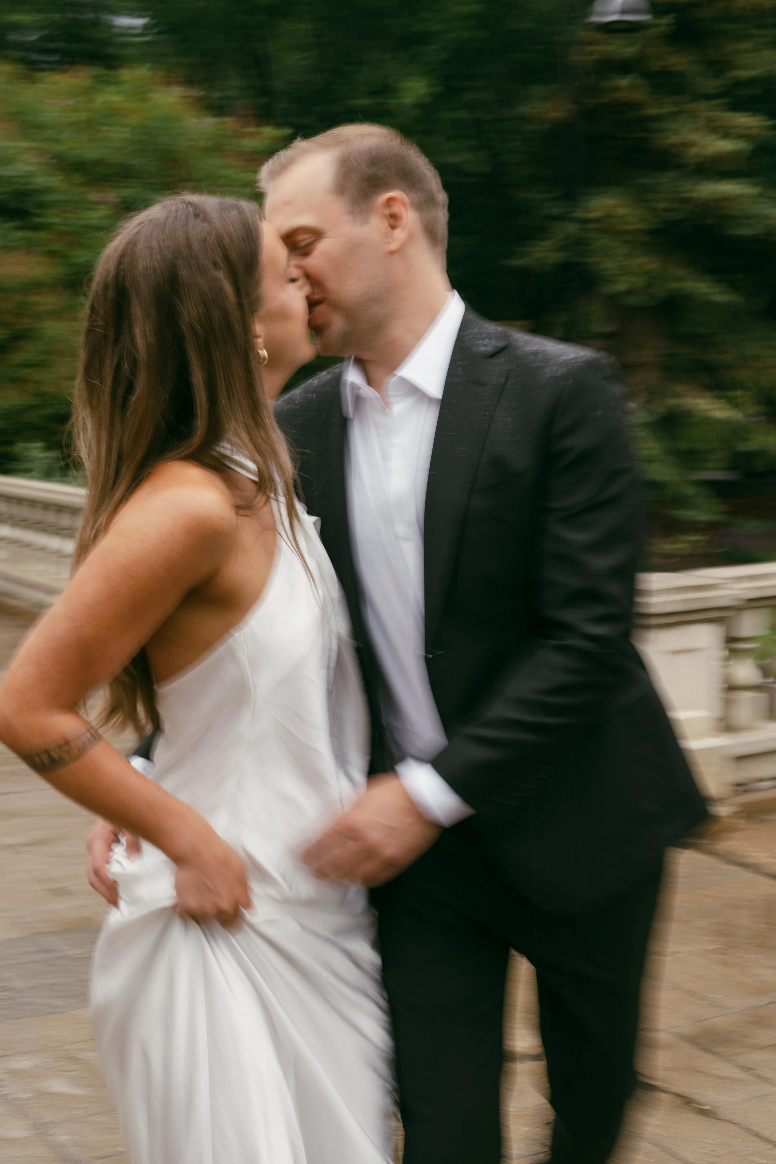 A couple in wedding attire sharing a kiss outdoors, with a blurred background of trees and a stone railing.