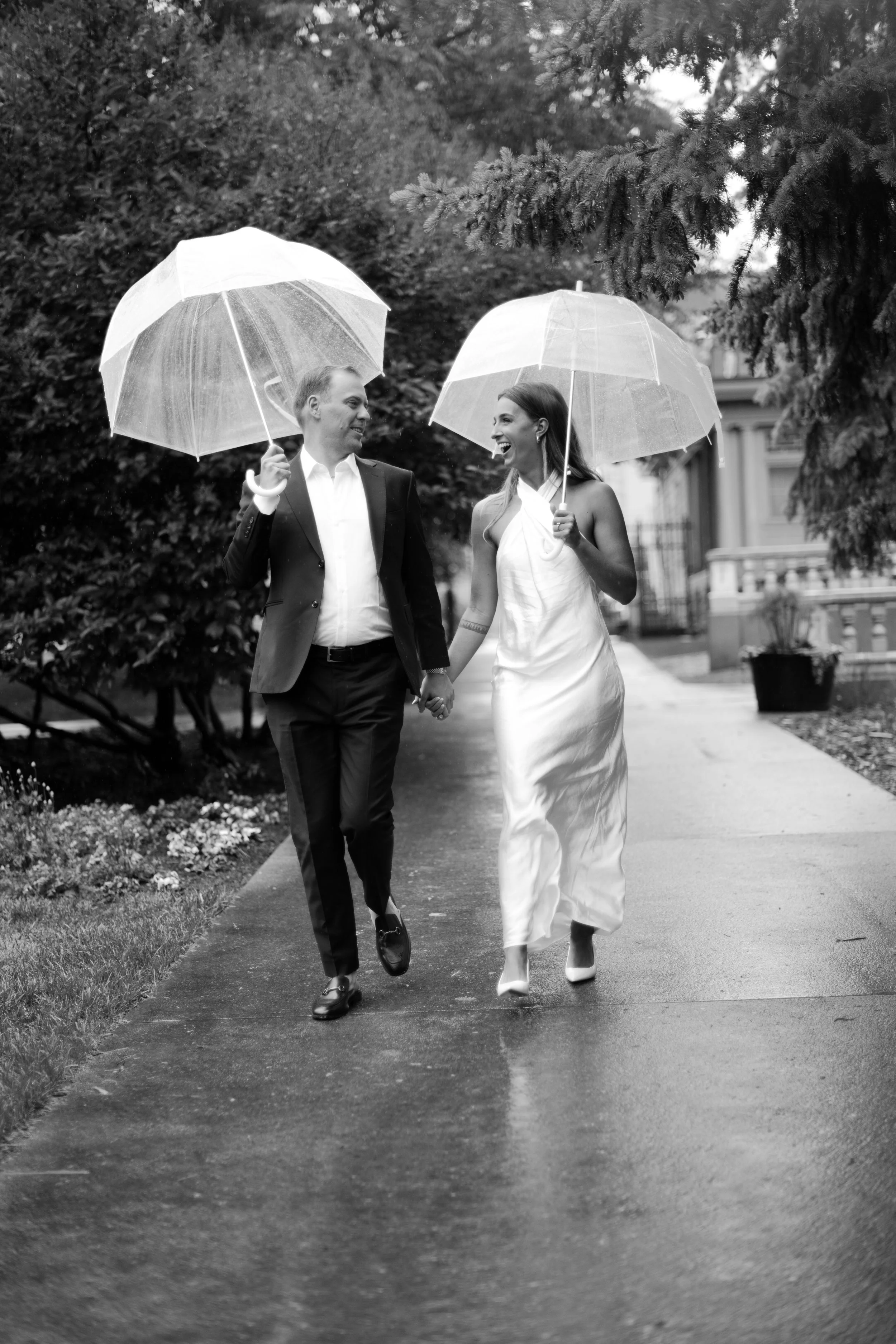 A couple dressed in formal attire walking hand in hand on a rainy day, holding umbrellas, smiling, and looking at each other.