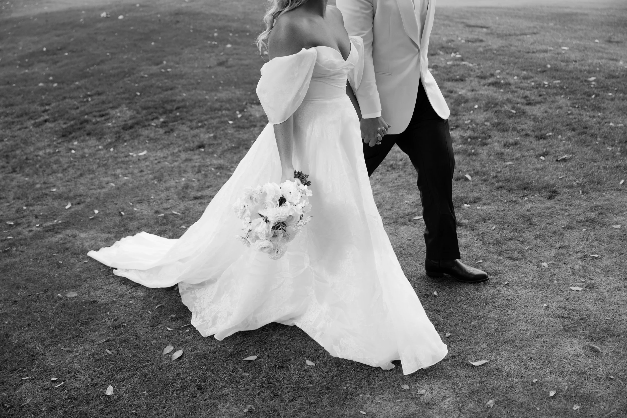 A black and white photo of a bride and groom holding hands outdoors on grass; the bride is wearing an off-the-shoulder wedding dress and holding a bouquet, the groom is in a light-colored jacket and dark pants.