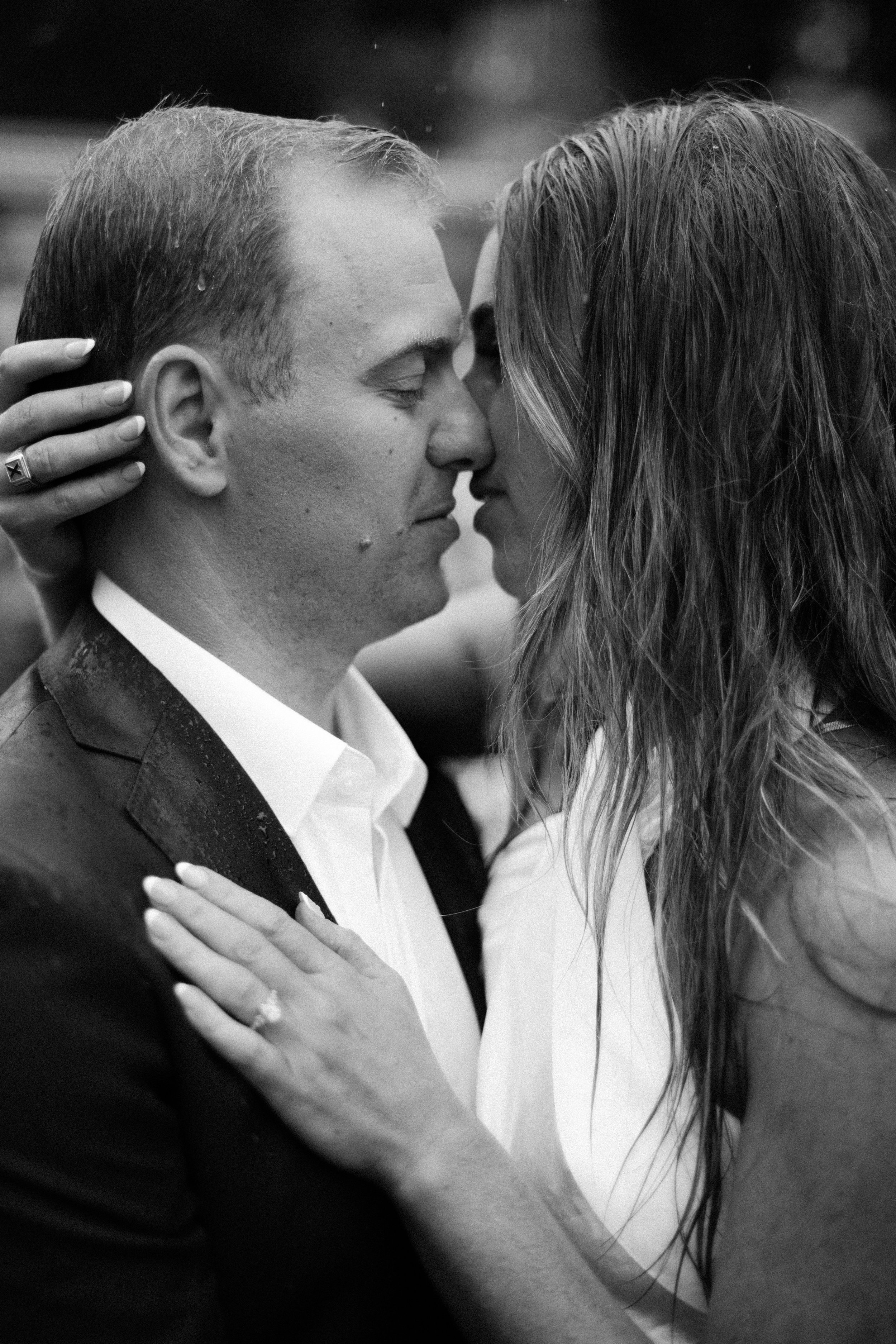 A black-and-white photo of a couple about to kiss, with their foreheads touching and eyes closed, outdoors in the rain.
