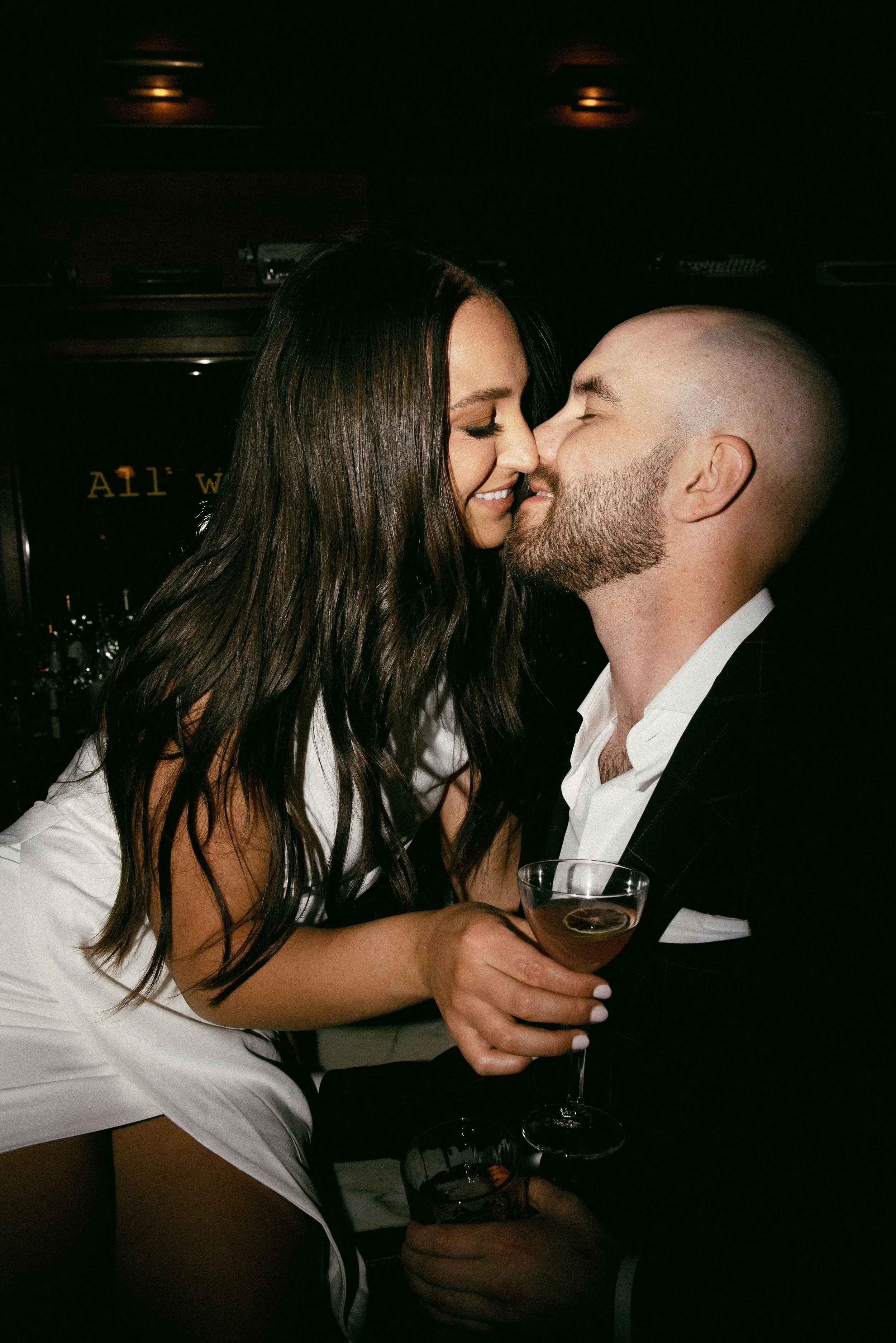 A woman and a man are close together, smiling and about to kiss in a dimly lit bar or nightclub. The woman has long dark hair and is wearing a white outfit, while the man has a bald head, a beard, and is dressed in a black suit and white shirt. They 