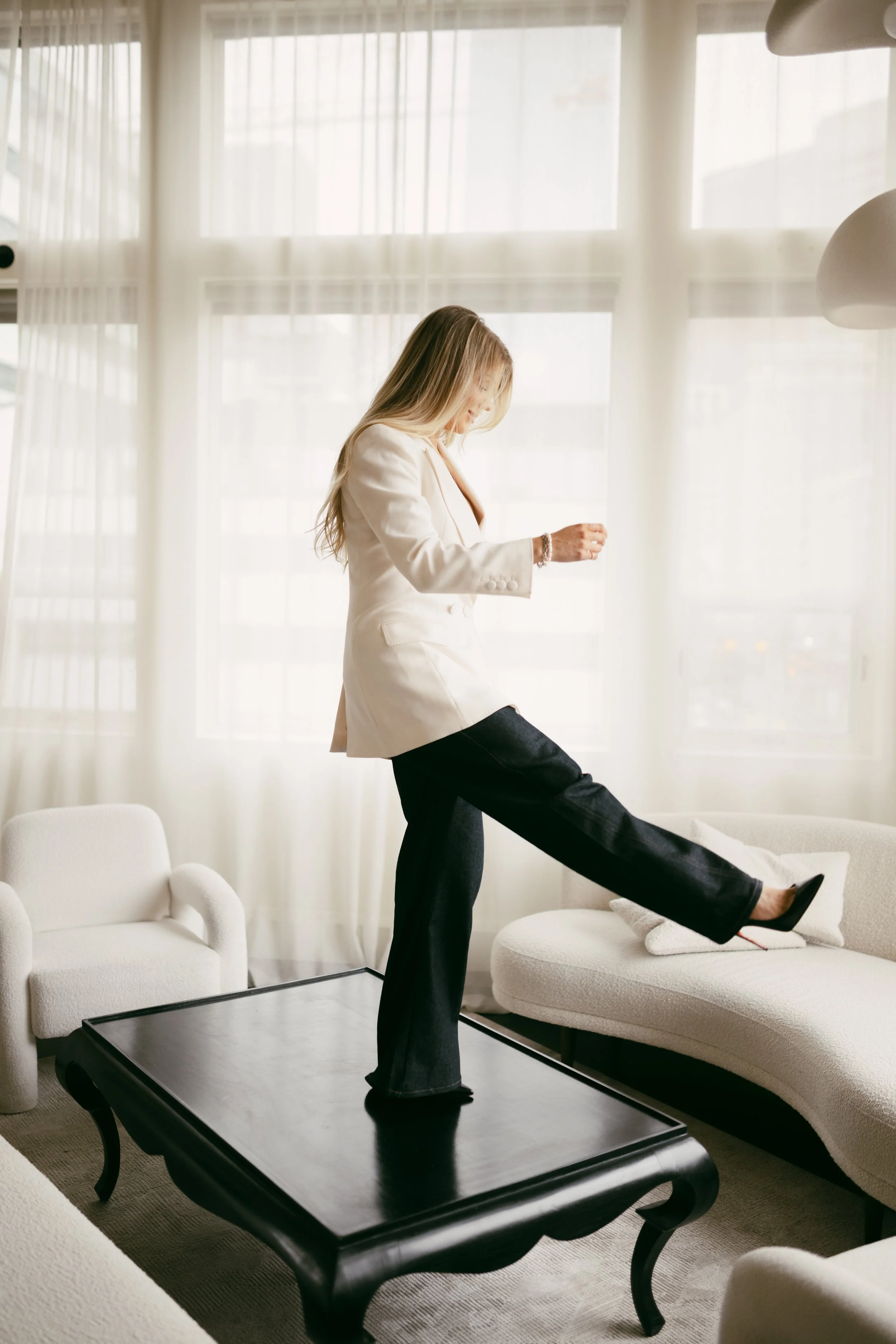 A woman in a white blazer and black pants standing on a black coffee table with one leg extended onto a white couch in a bright living room with large windows and sheer curtains.