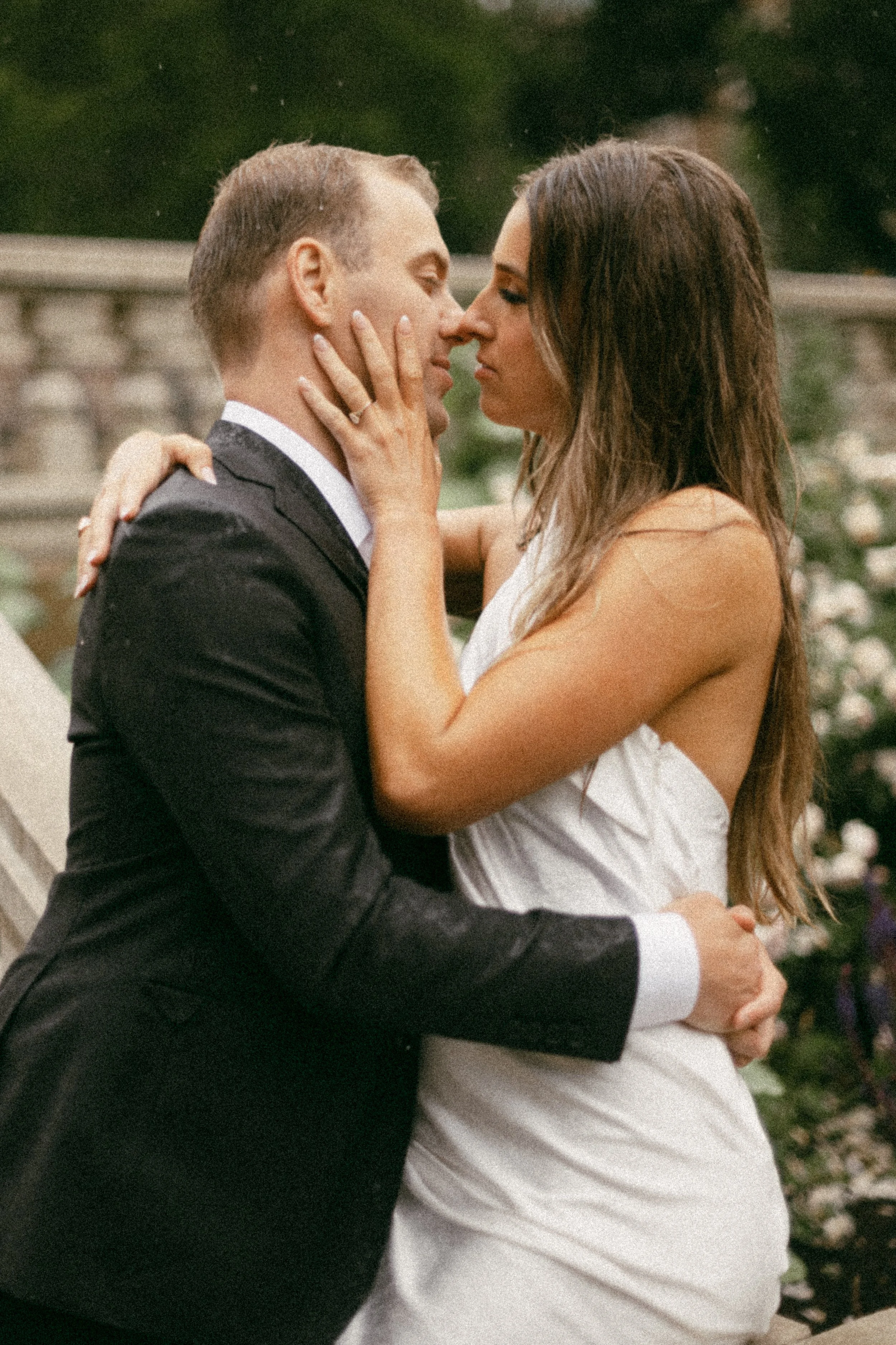 A man in a black tuxedo and a woman in a white dress embrace each other intimately outdoors, with their faces close and eyes closed, in a romantic moment.