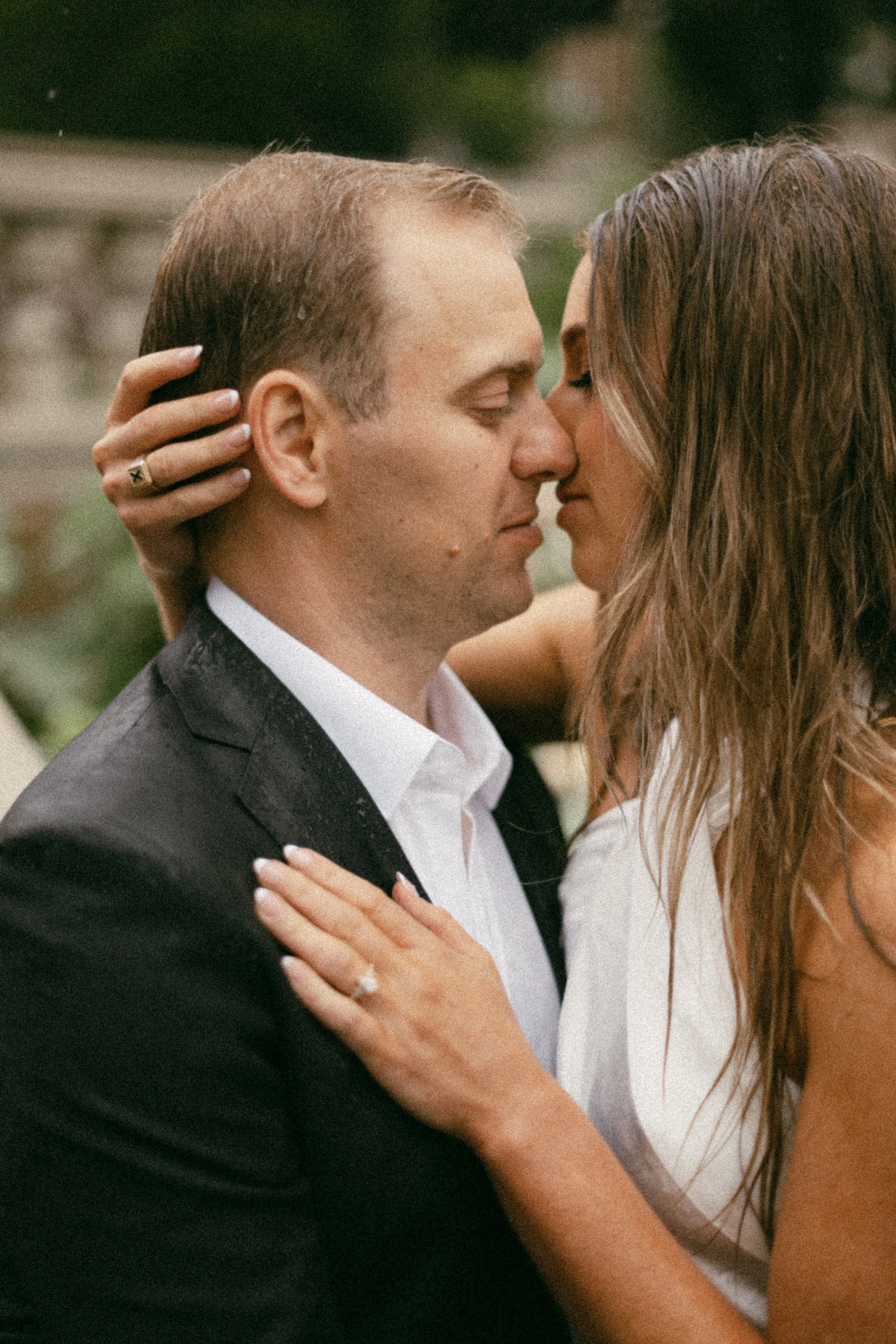 A man and woman are close together, with their foreheads and noses touching in an intimate moment, outdoors. The woman has her hand on the man's shoulder, and the man is dressed in a black suit and white shirt. The woman has long wavy hair and wears 