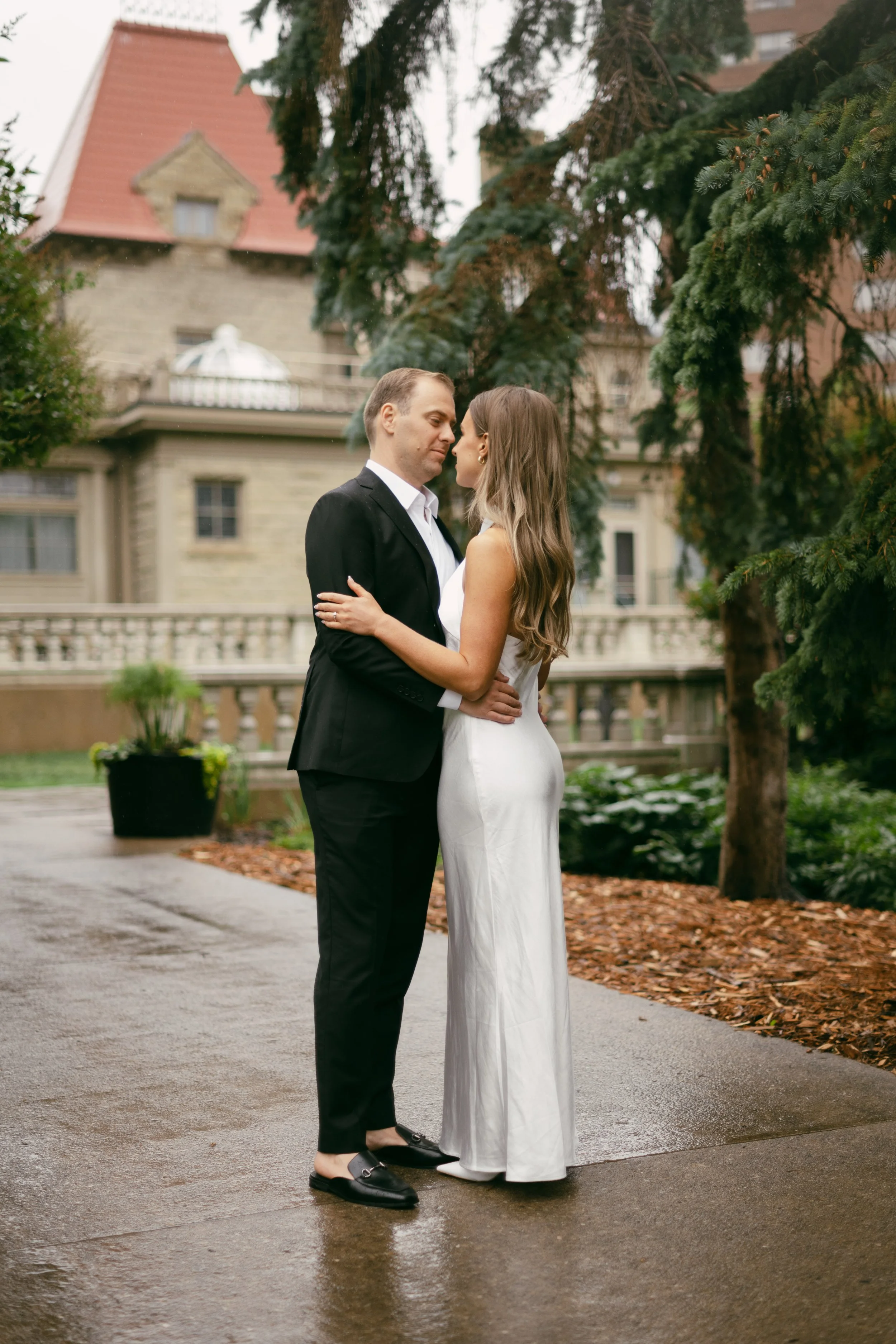 A couple in formal attire standing close together outdoors, looking at each other with a mansion and large trees in the background.