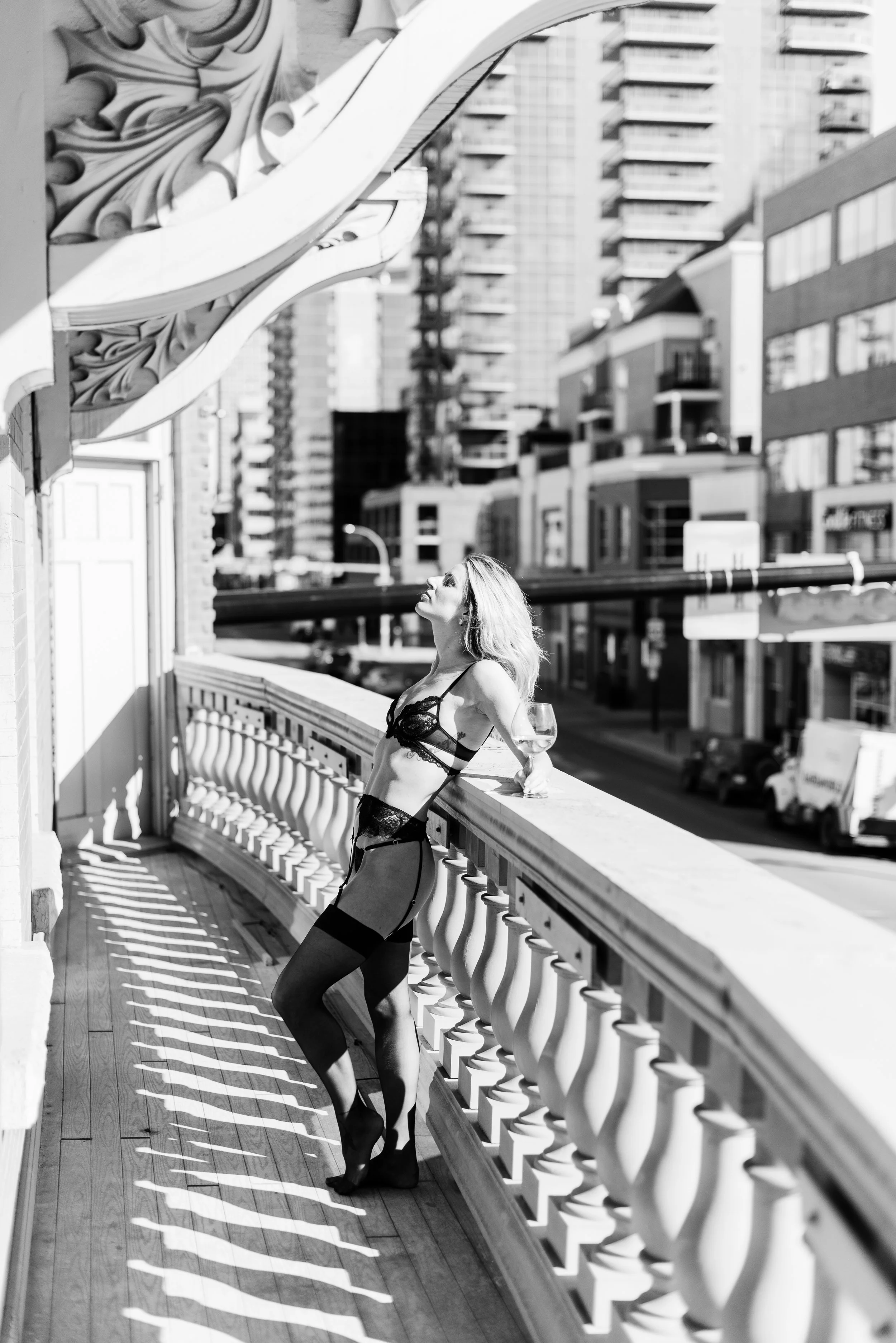 A woman in lingerie and stockings leaning on a balcony railing with a glass of wine, looking upward in an urban cityscape with tall buildings.