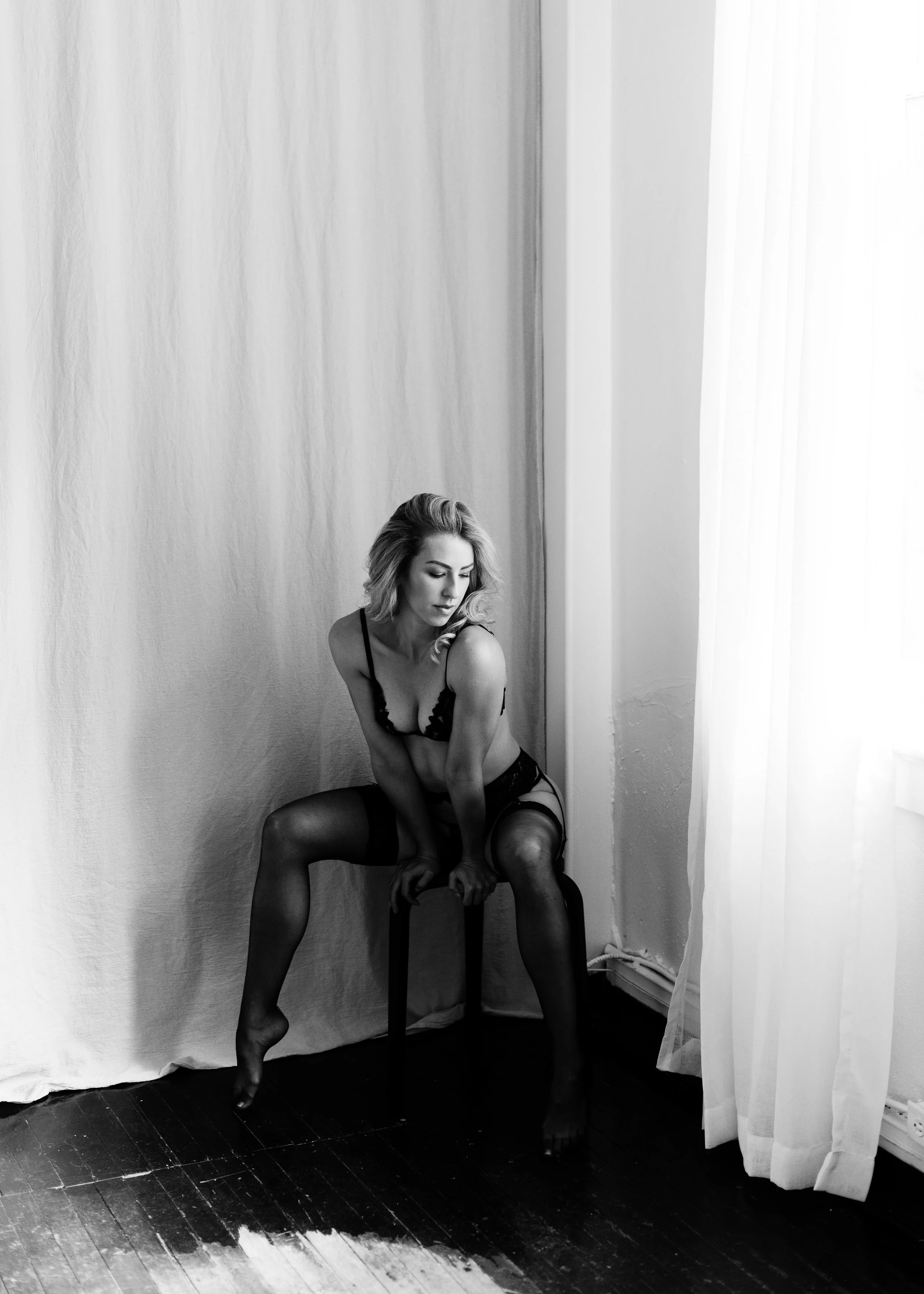 A woman with blonde hair wearing black lingerie and stockings, sitting on a chair in a room with white curtains and a textured wall, looking down to her left.