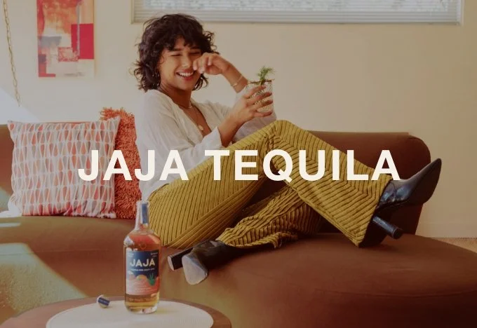 A woman sitting on a couch holding a small plant in a glass, smiling and laughing, with a bottle of tequila labeled 'Jaja' on a table in front of her, indoor setting with a window and artwork in the background.