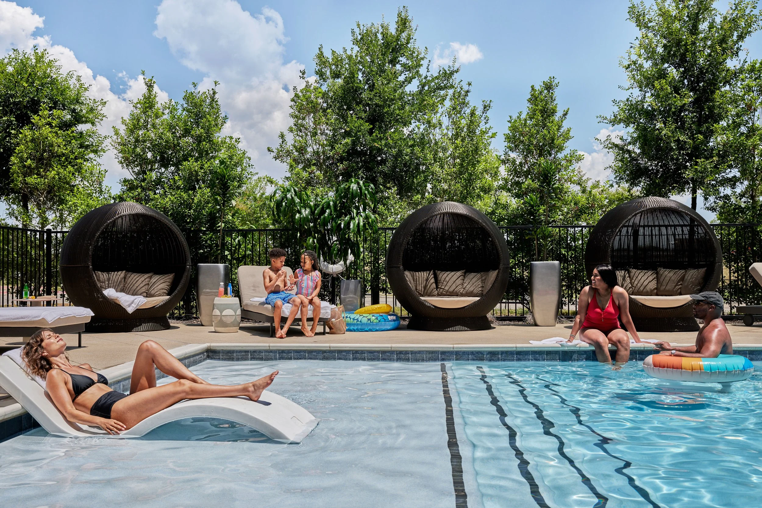 People relaxing by a swimming pool on a sunny day, with some sitting on poolside loungers, a woman sunbathing on a lounge chair, two children sitting on a poolside bench, and a woman and man sitting in the pool with pool toys, surrounded by trees and