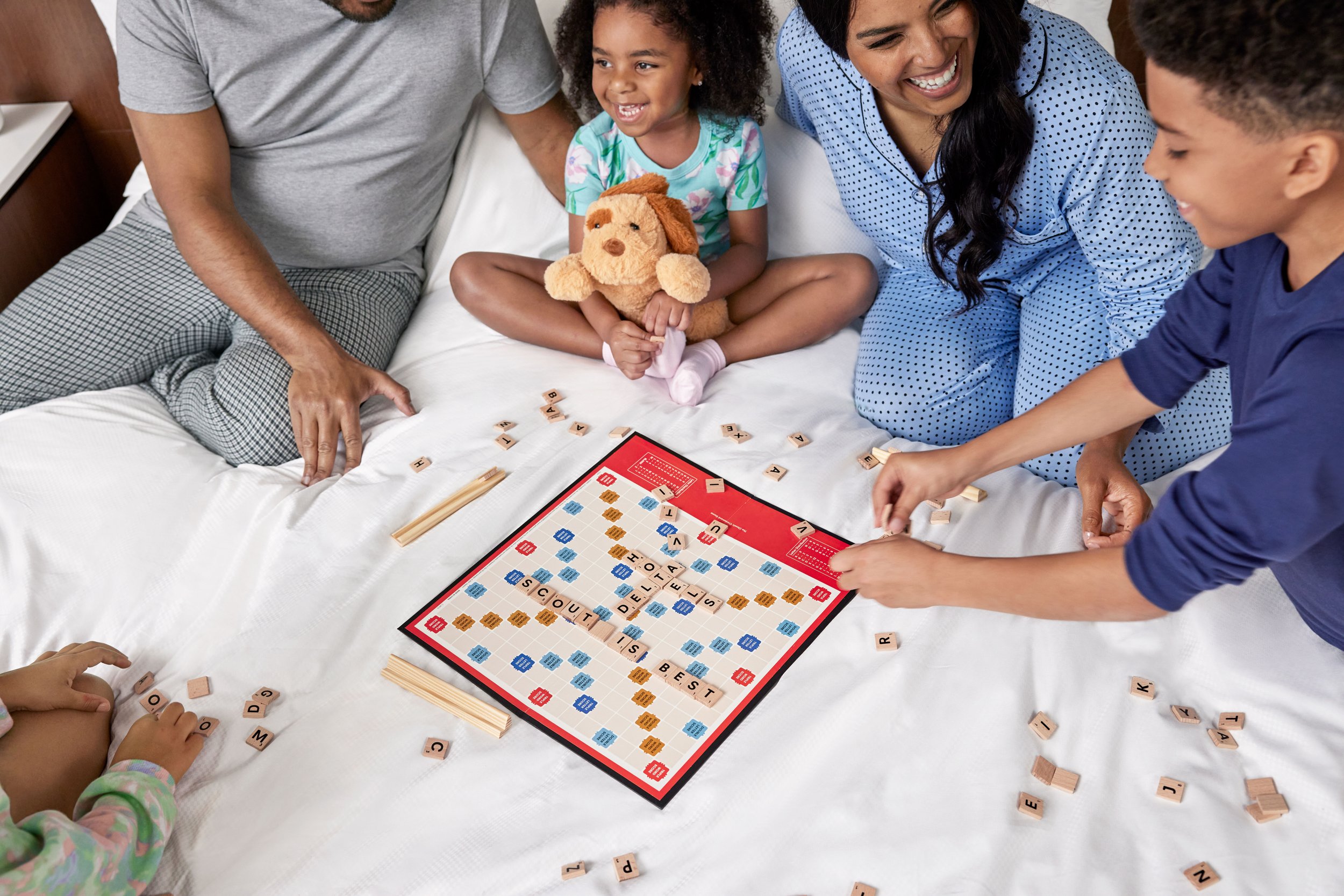 A family is playing Scrabble on a bed, with a young girl holding a teddy bear and smiling, while a woman and a boy are engaged in the game, with letter tiles scattered around.