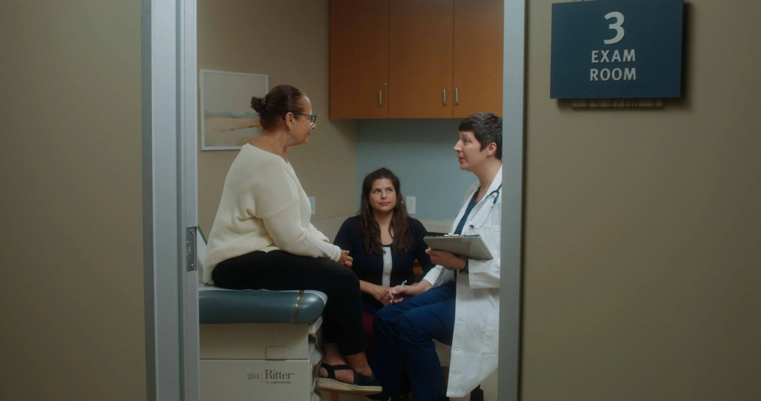 Doctor speaking with two women in an exam room labeled '3'. One woman is seated on the examination table, and the other woman and the doctor are standing and holding hands.