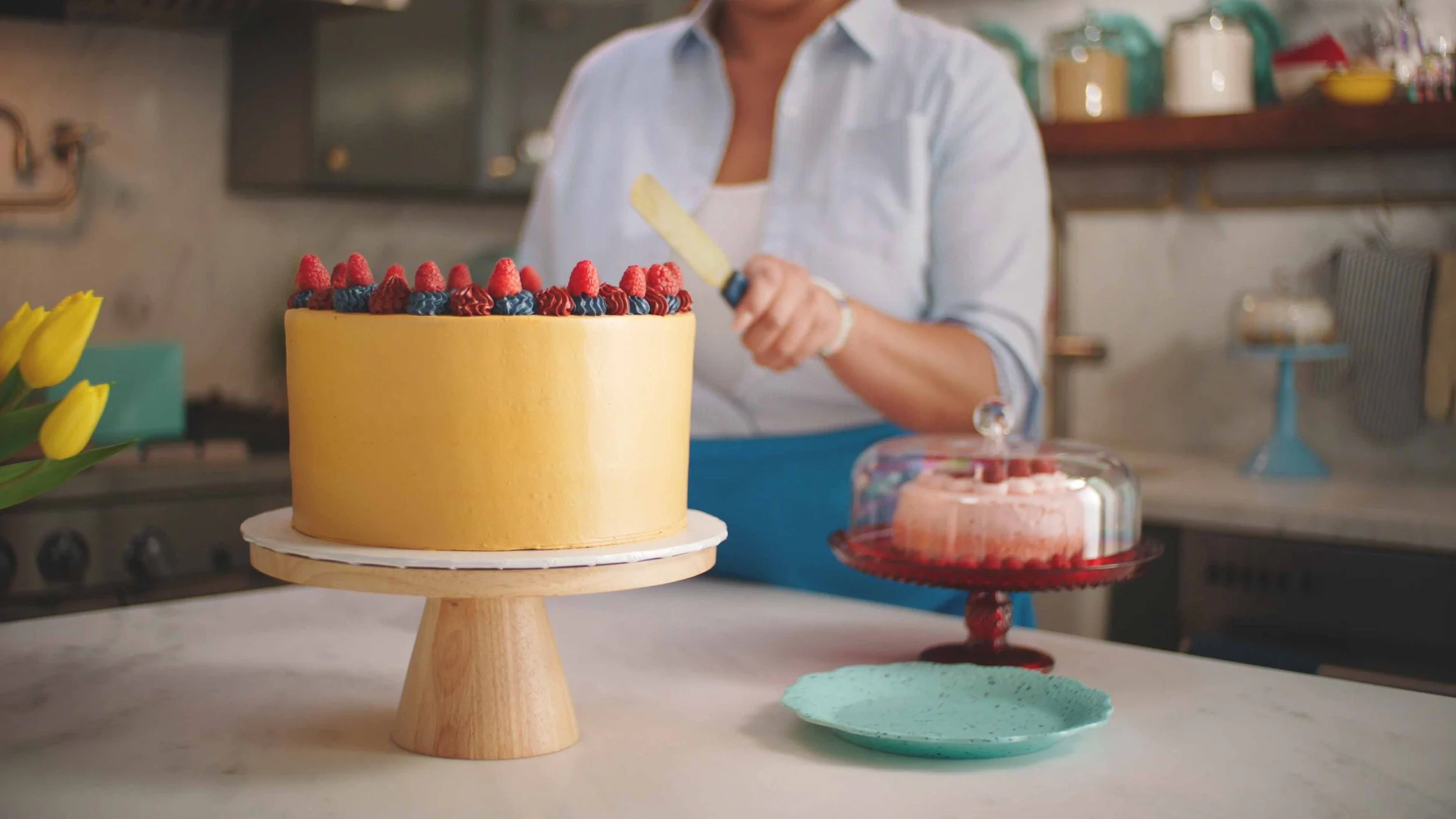 Woman holding a knife on a kitchen countertop with a yellow frosted cake topped with raspberries and blueberries, next to a pink cake under a glass dome, and a turquoise plate.
