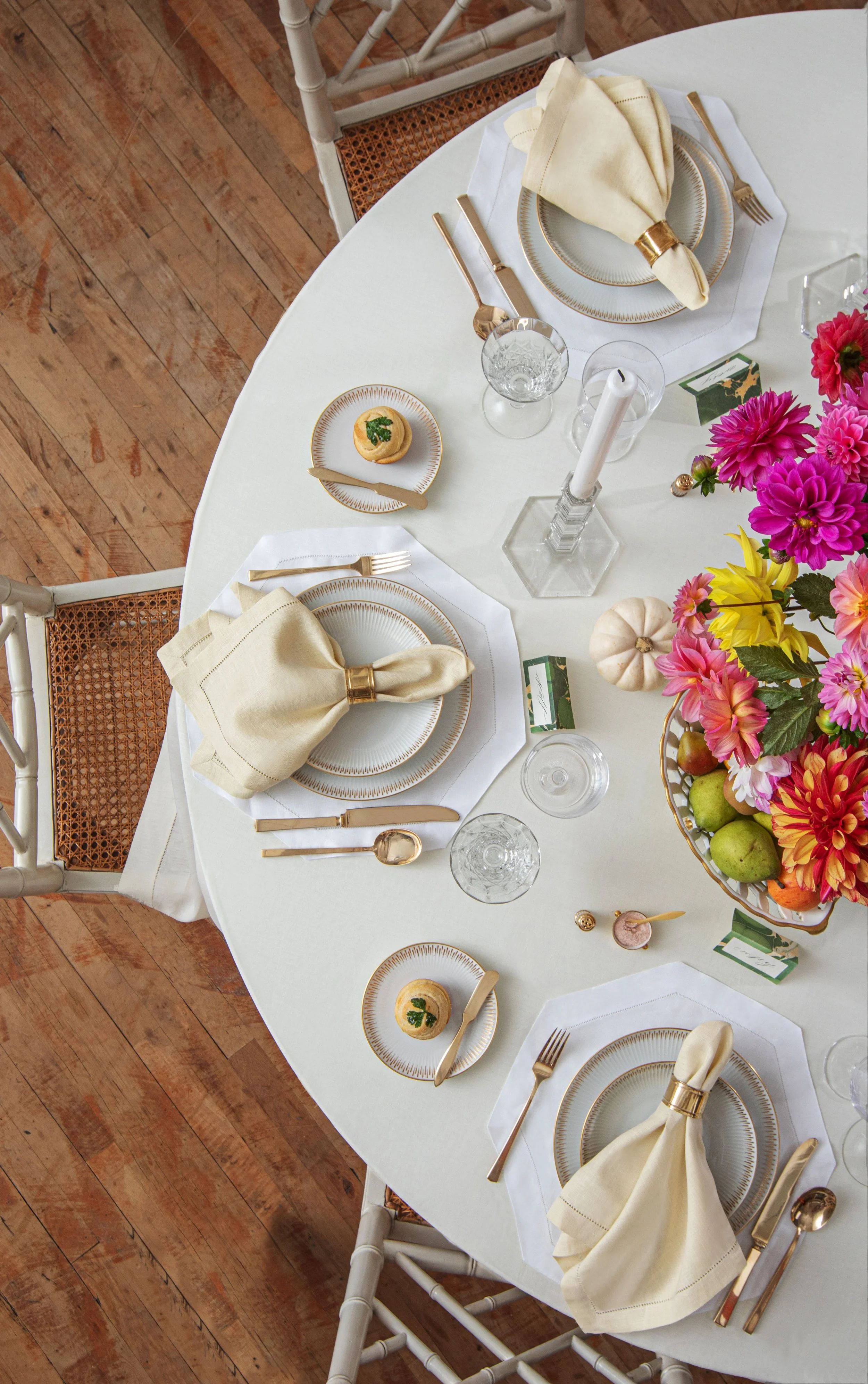 Round dining table set with white tablecloth, gold-rimmed white and gold dinnerware, gold flatware, cream-colored napkins with gold napkin rings, clear glassware, and a centerpiece of pink, purple, and yellow flowers in a gold bowl. Small decorative pumpkins and greenery accents add a festive touch.