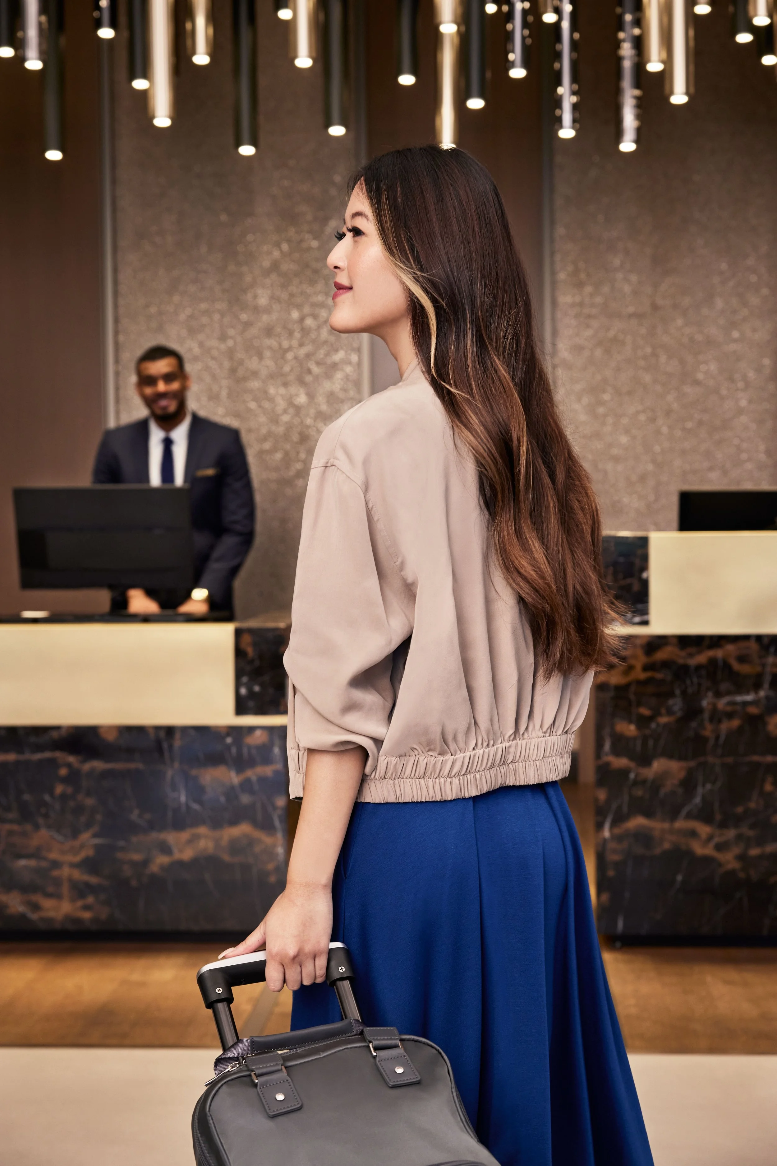 A woman with long brown hair wearing a beige jacket and blue skirt waiting at a hotel reception with her rolling suitcase, while a man in a suit smiles behind the counter.