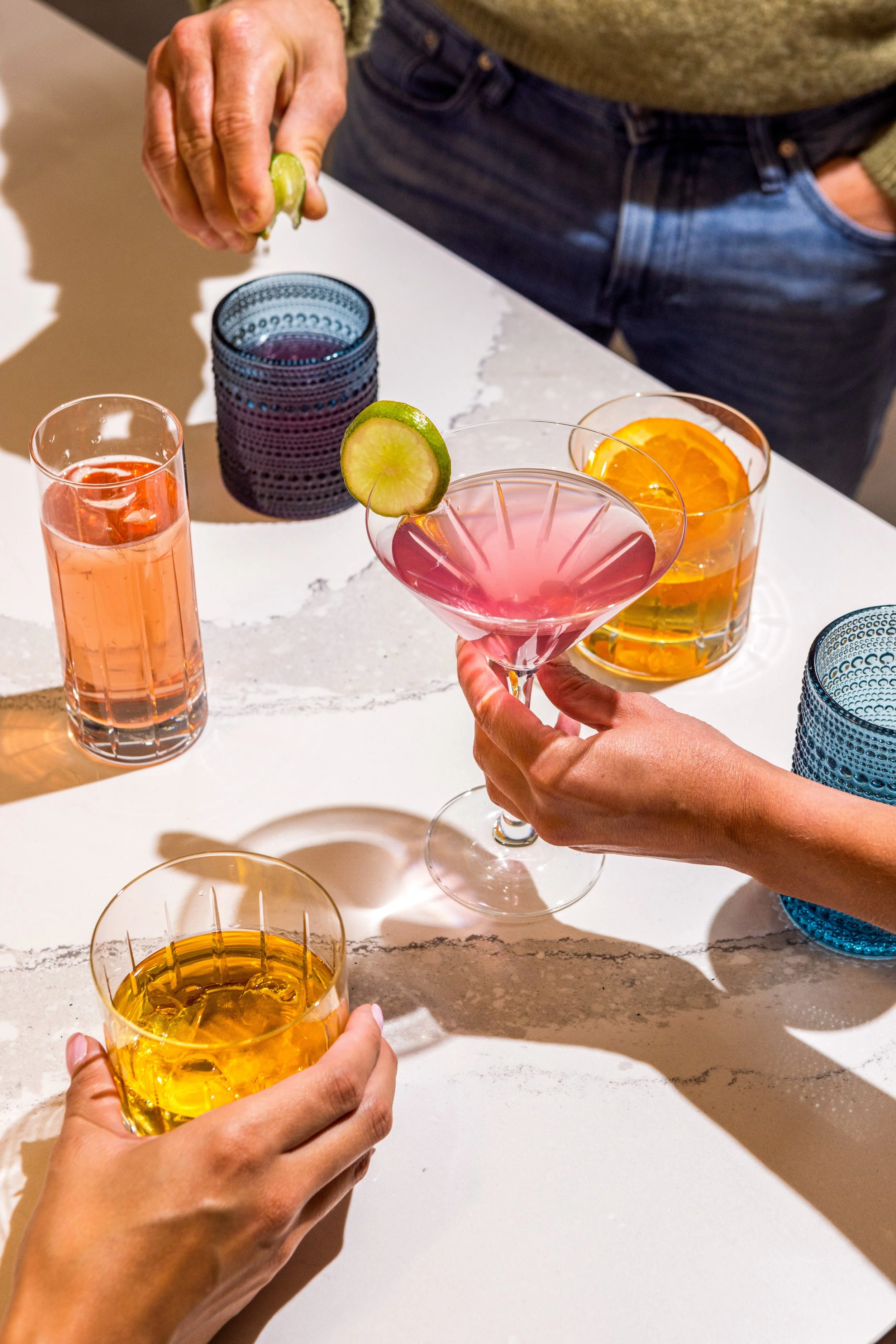People holding colorful cocktails and glasses on a table at a social gathering.