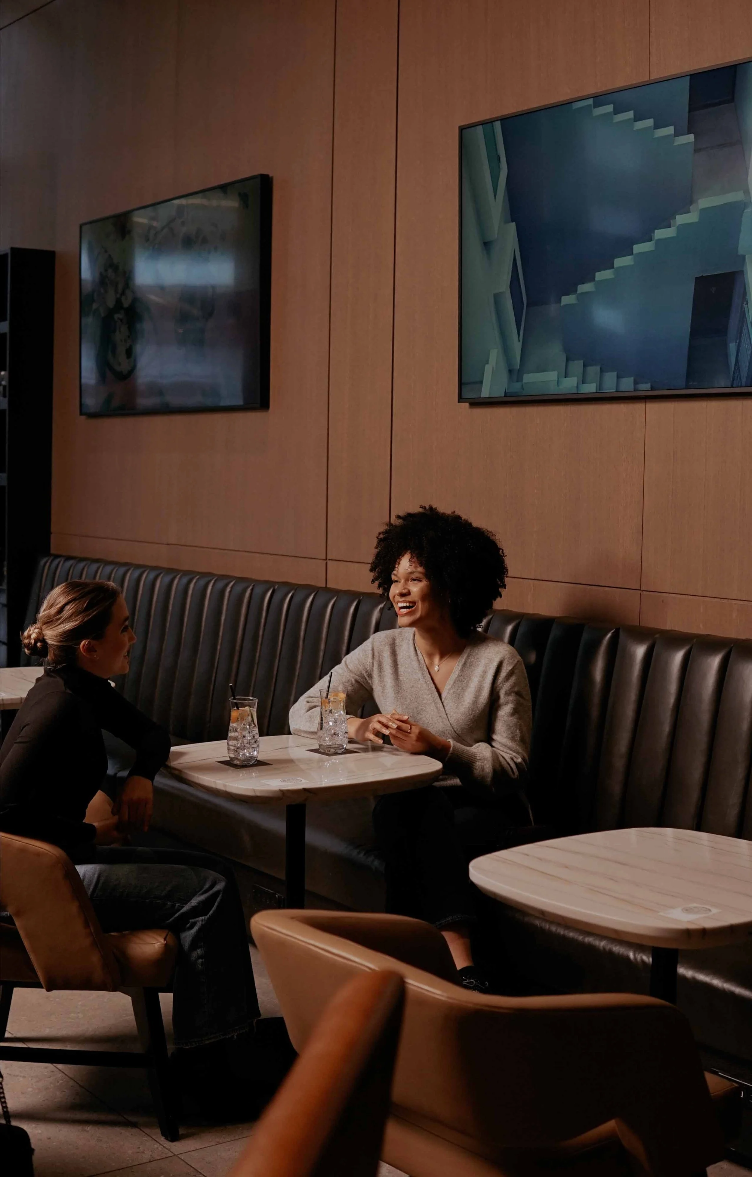 Two women sitting across from each other at a restaurant table, smiling and talking. The woman on the left has brown hair pulled back, wearing black, and the woman on the right has curly black hair, wearing a beige sweater. There are glasses with drinks on the table, and large screens on the wood-paneled wall behind them.