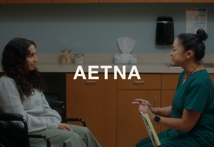 A healthcare professional in green scrubs speaking with a young female patient in a medical office, with the text 'AETNA' overlaid.