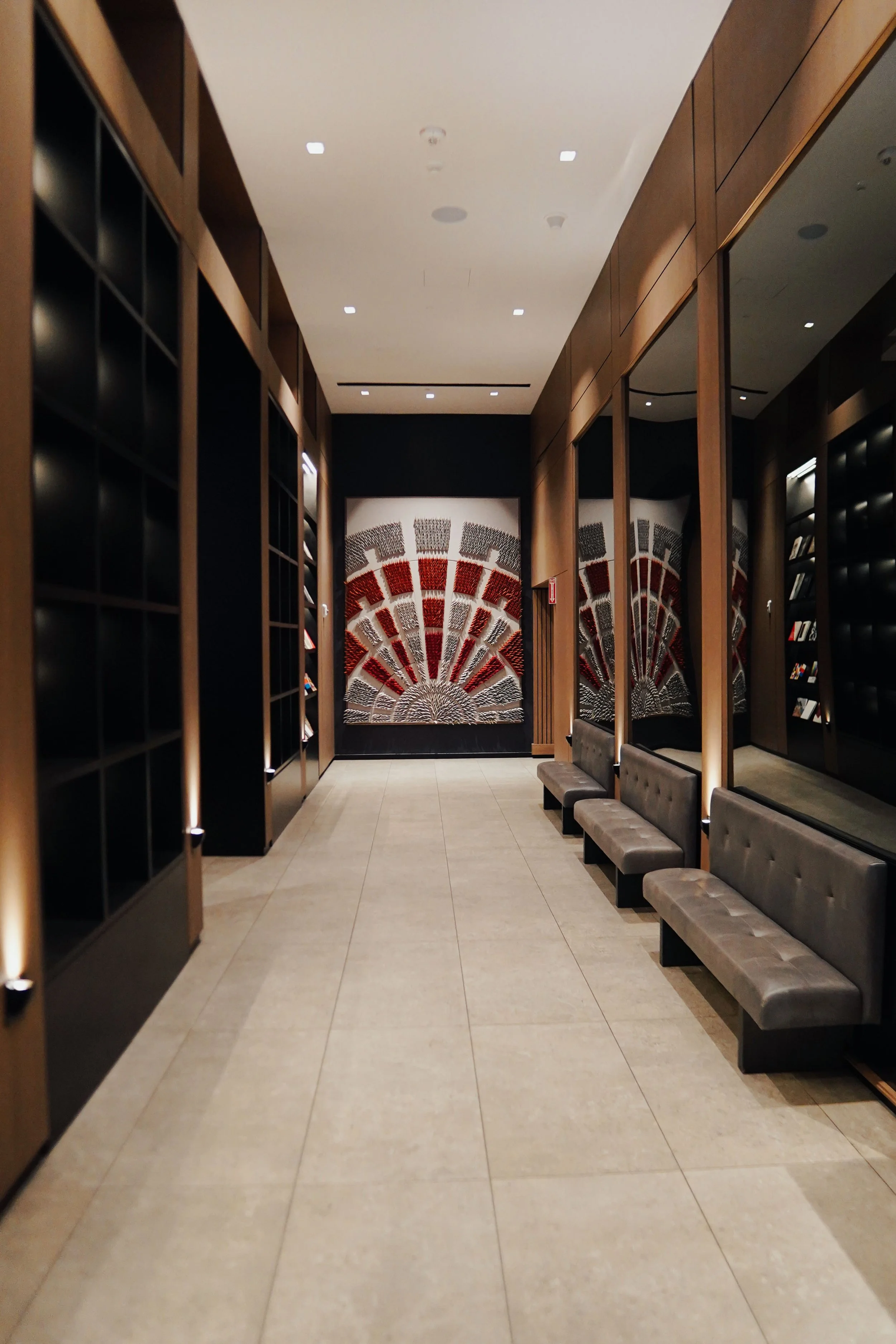 Modern hotel hallway with beige tile floors, wooden-paneled walls, and black shelving units. There are four gray cushioned benches along the wall and abstract artwork at the end of the corridor.