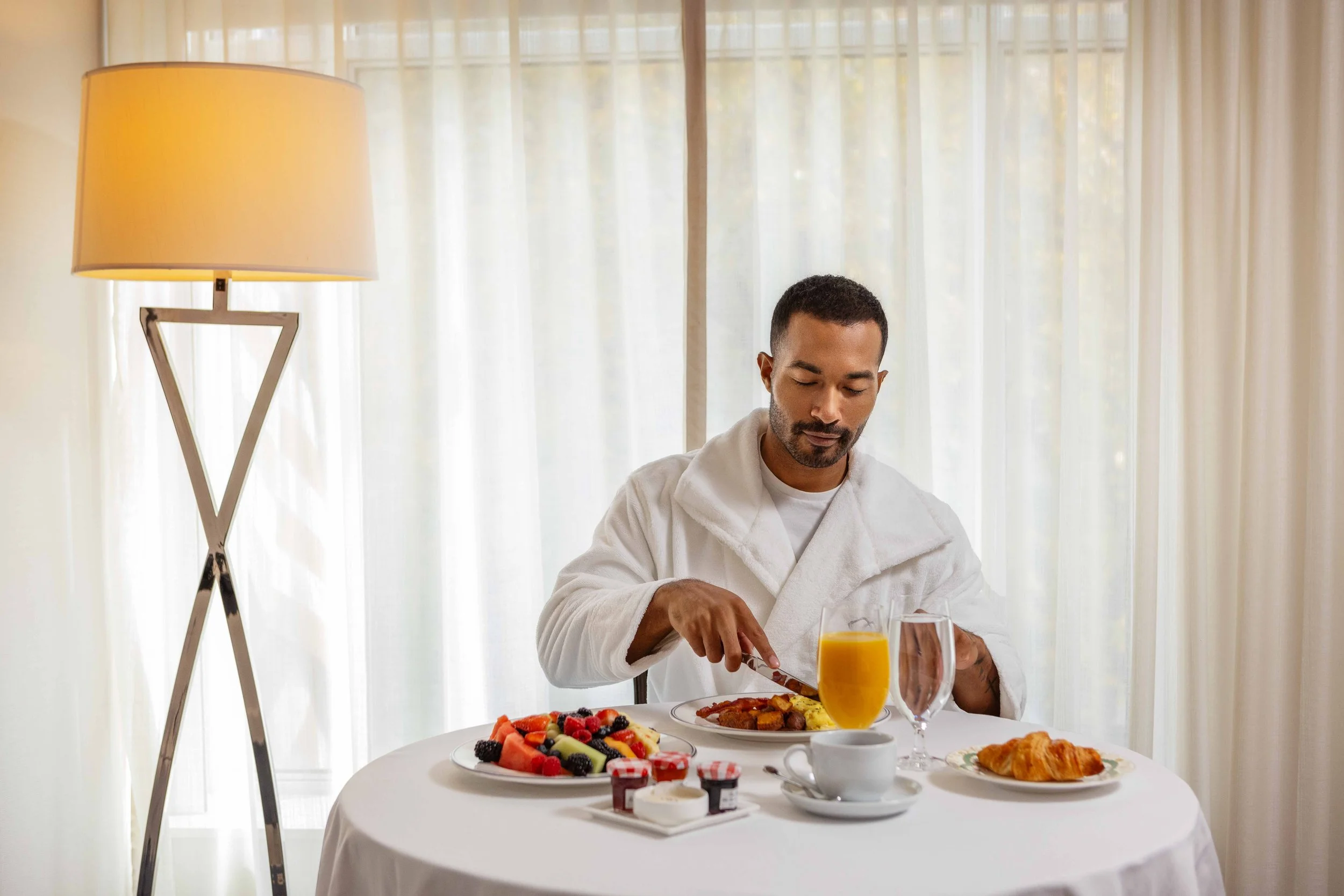 Man in a white robe eating breakfast at a table with fruit, scrambled eggs, croissant, juice, coffee, and jam, in a bright room with curtains.