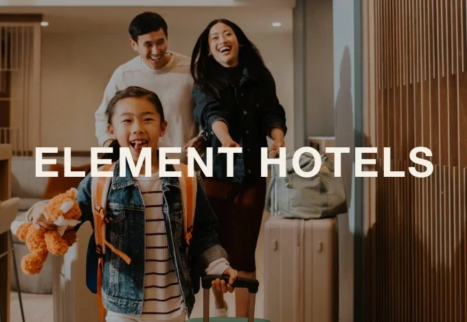 A family of three, including a man, a woman, and a young girl, smiling and welcoming at a hotel lobby with luggage, promoting Element Hotels.