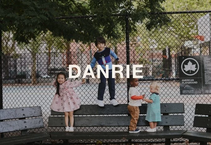 Four young children playing and interacting on a park bench in front of a chain-link fence at an outdoor playground.