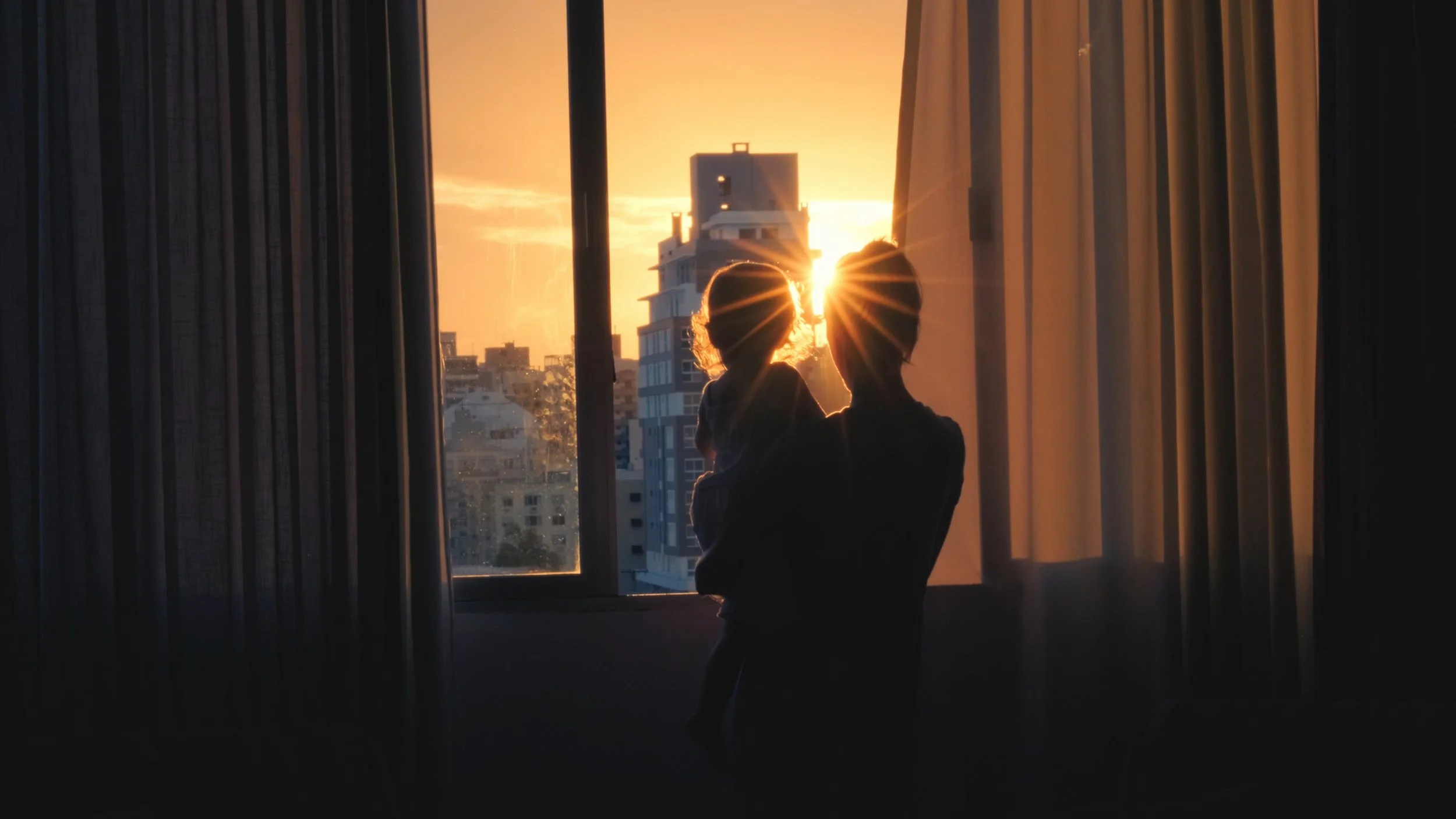 Person holding a child in front of a window at sunset, with city buildings outside.