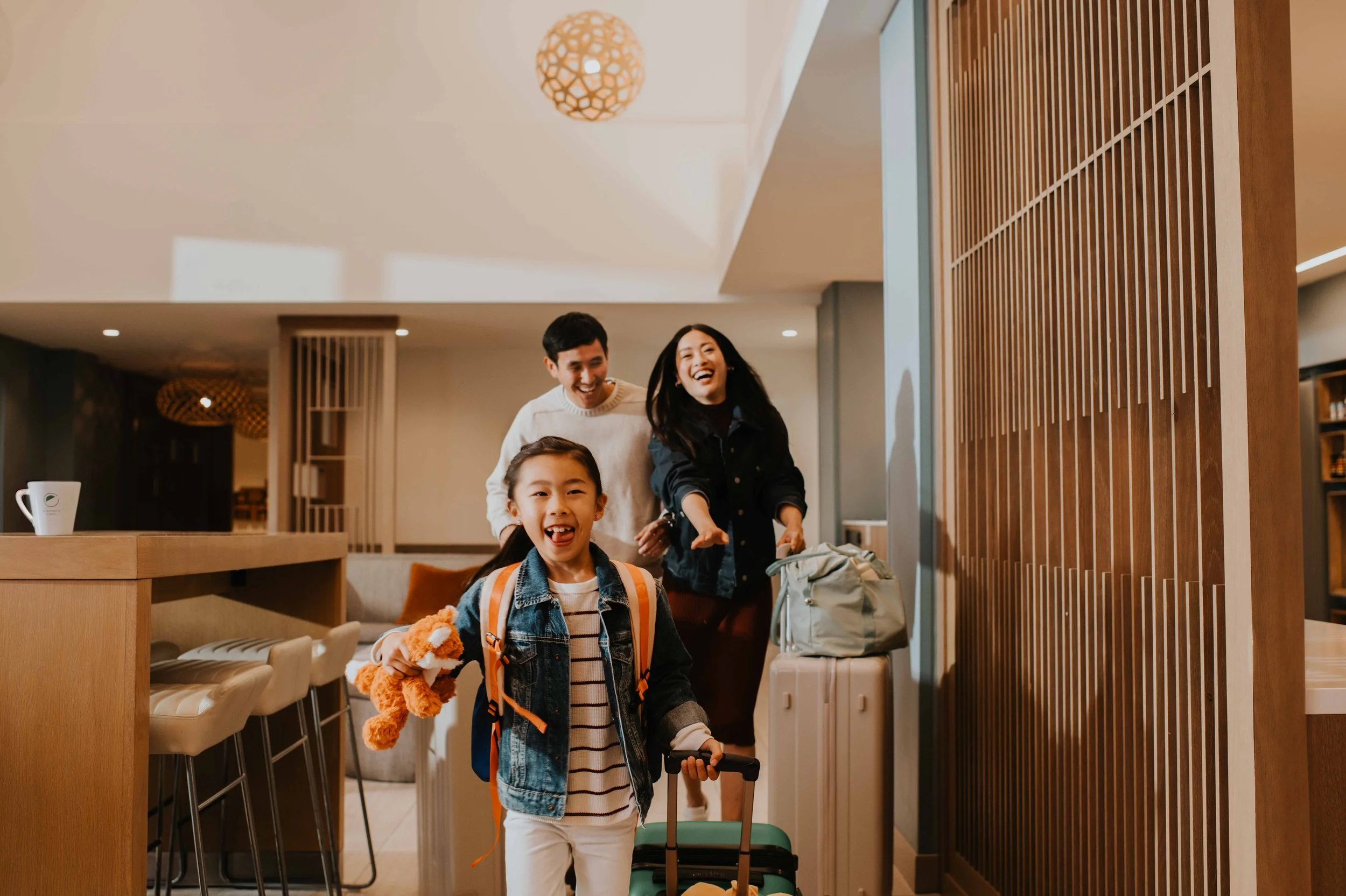 A family of three arriving at a hotel, smiling and happy. The young girl is in front with a backpack and stuffed animal, pulling a small suitcase. The parents are behind her, also smiling and happy.
