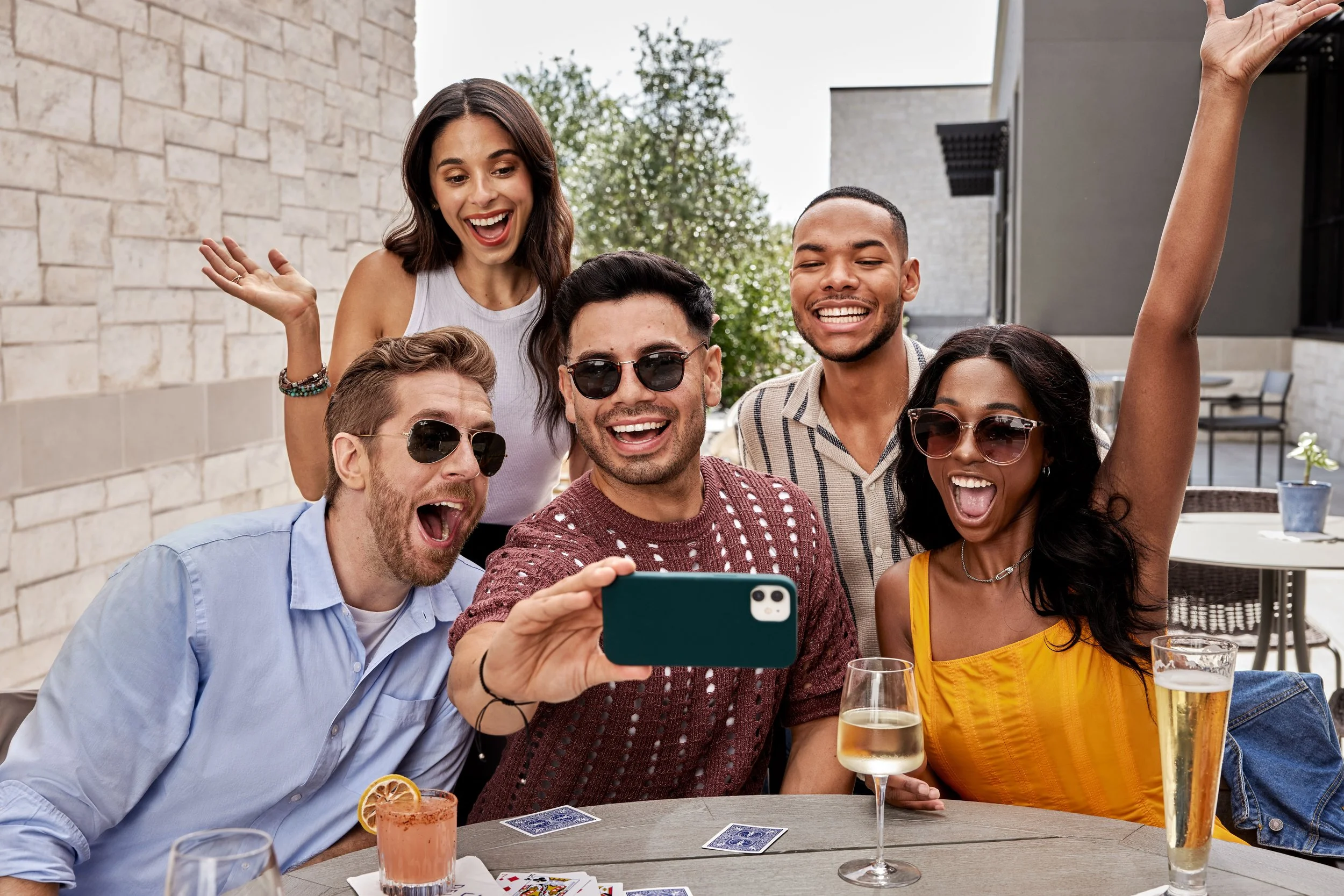 A group of six friends, three men and three women, are taking a selfie together at an outdoor gathering. They are smiling and wearing sunglasses, with drinks on the table in front of them.