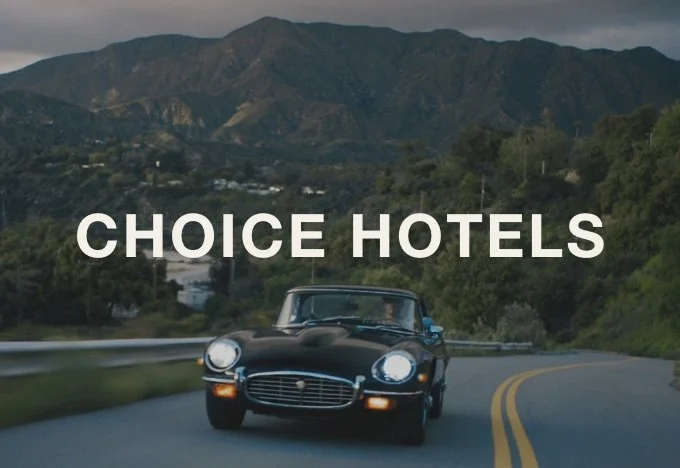 A black vintage car driving on a winding mountain road with green hills and mountains in the background, and the text 'CHOICE HOTELS' superimposed.