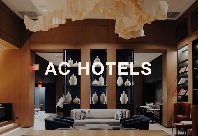 Hotel lobby with a large wooden wall display of decorative vases, a gray sofa, black armchairs, and a bookshelf on the right.