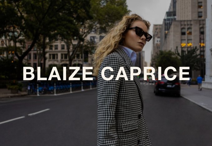 A woman with curly blonde hair wearing sunglasses, a plaid blazer, and a blue shirt standing on a city street with buildings, trees, and cars in the background.